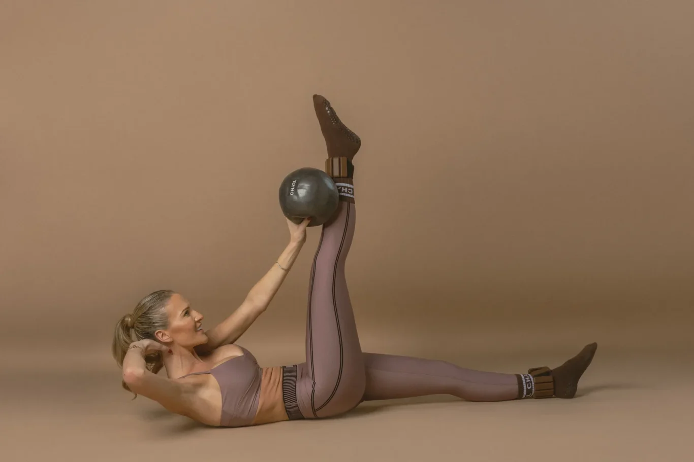 Athlete in mauve athletic wear lying on her back with legs raised, holding a kettlebell between her legs against a brown studio backdrop.