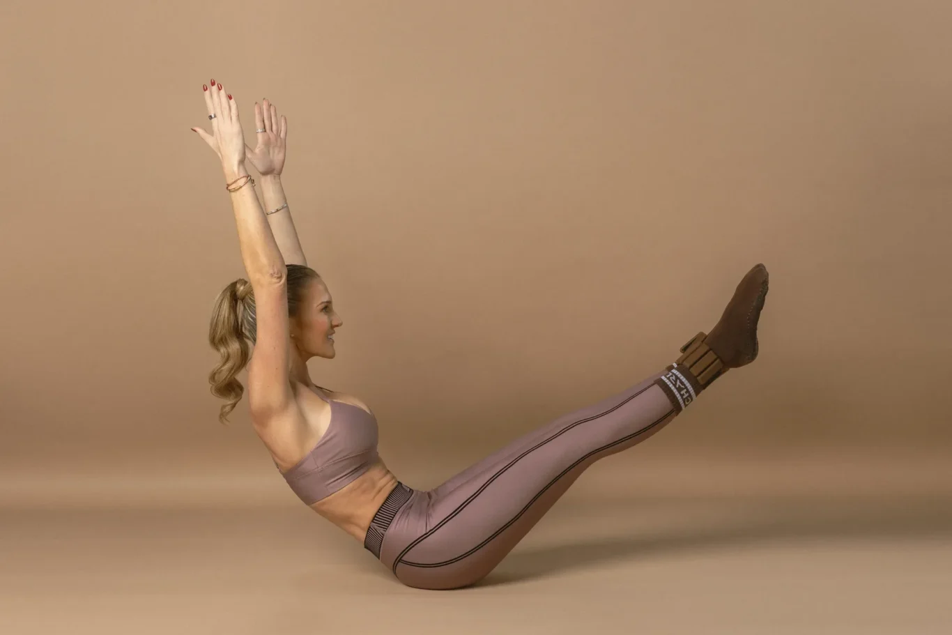 Woman in pink athletic wear on the floor, arms raised overhead as she performs a high leg raise with legs extended into a long split against a beige background.