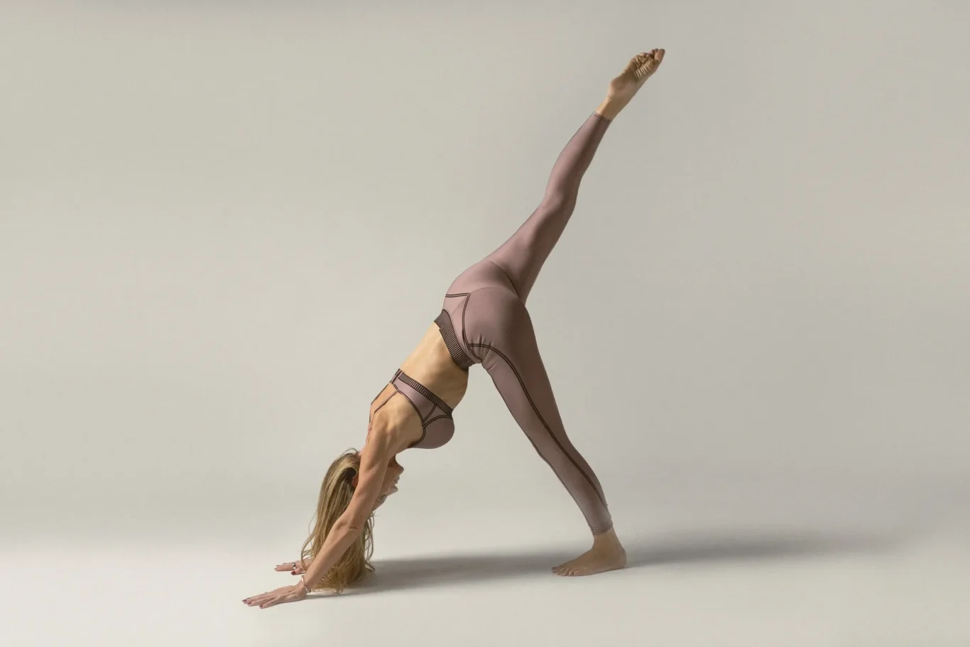 Person in mauve athleticwear balances on their hands with one leg extended straight up in a yoga pose on a light floor surface.