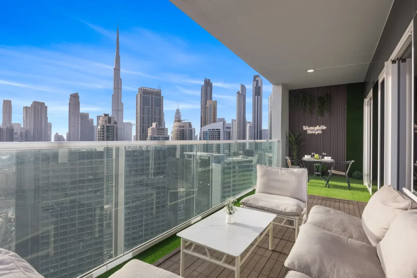 Balcony with white cushioned seating, a white coffee table, and glass railing overlooking a blue-sky city skyline.
