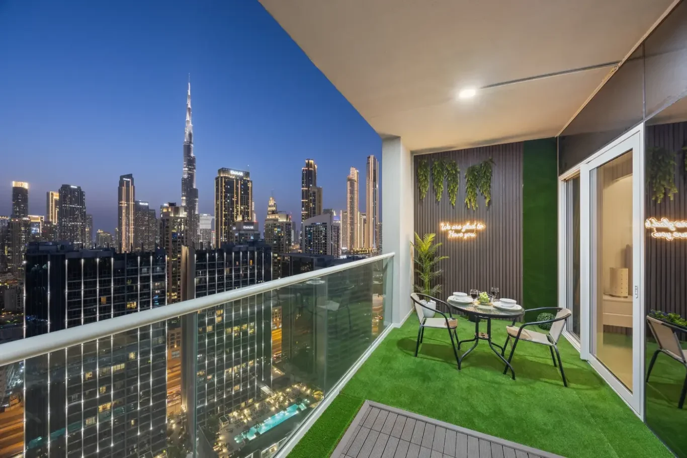 Night skyline of illuminated skyscrapers viewed from a glass-railed balcony with a small round dining set on green artificial turf.
