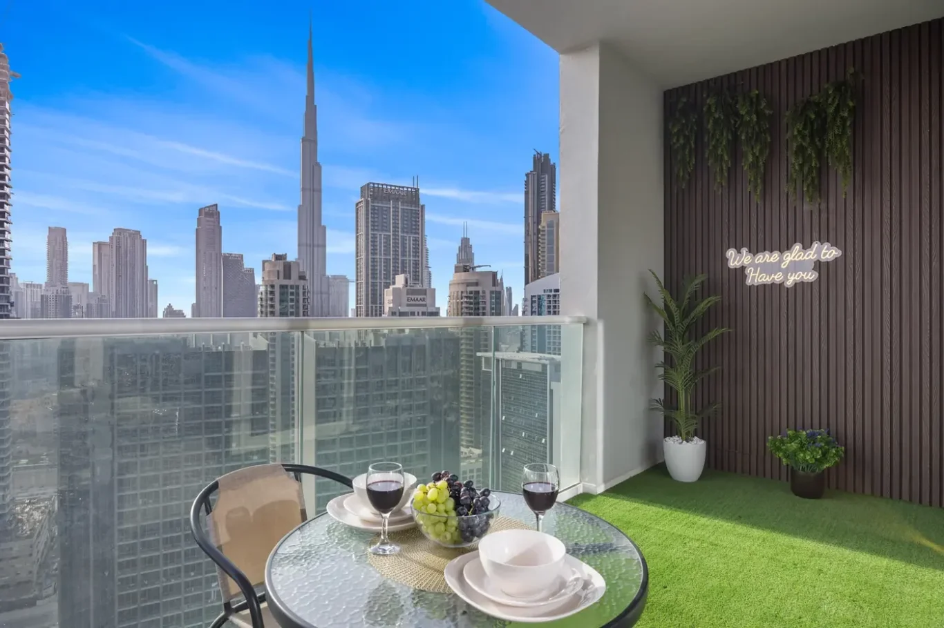 Balcony dining table with two wine glasses and fruit bowl, overlooking a city skyline with tall skyscrapers.