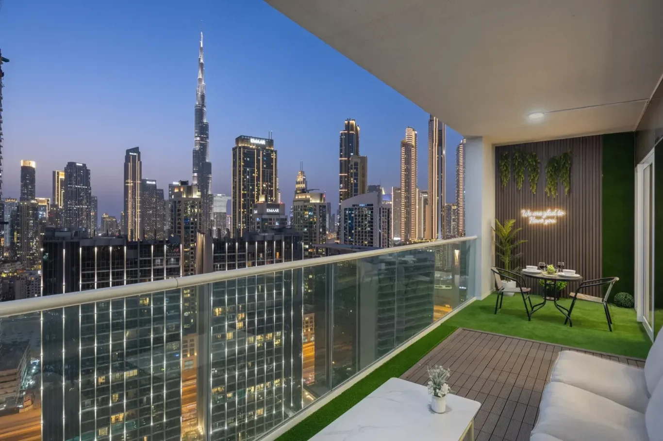 Balcony at dusk overlooking a lit city skyline; glass railing, white outdoor seating, a small table with plants, and a round dining table on artificial turf below with a neon sign on the wall.