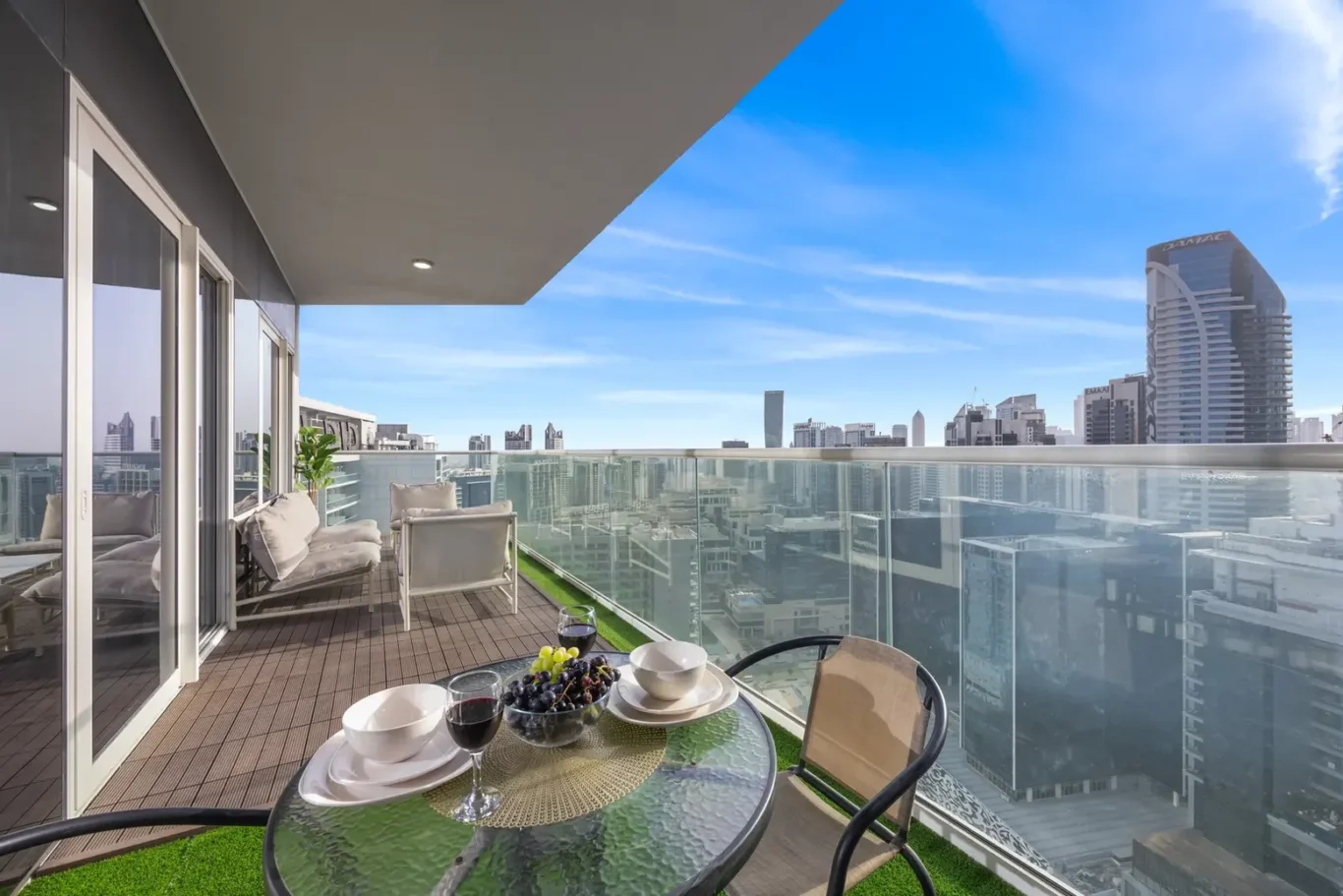 Balcony with glass railing overlooking a city skyline; table set for a meal with fruit, bowls, and wine glasses.