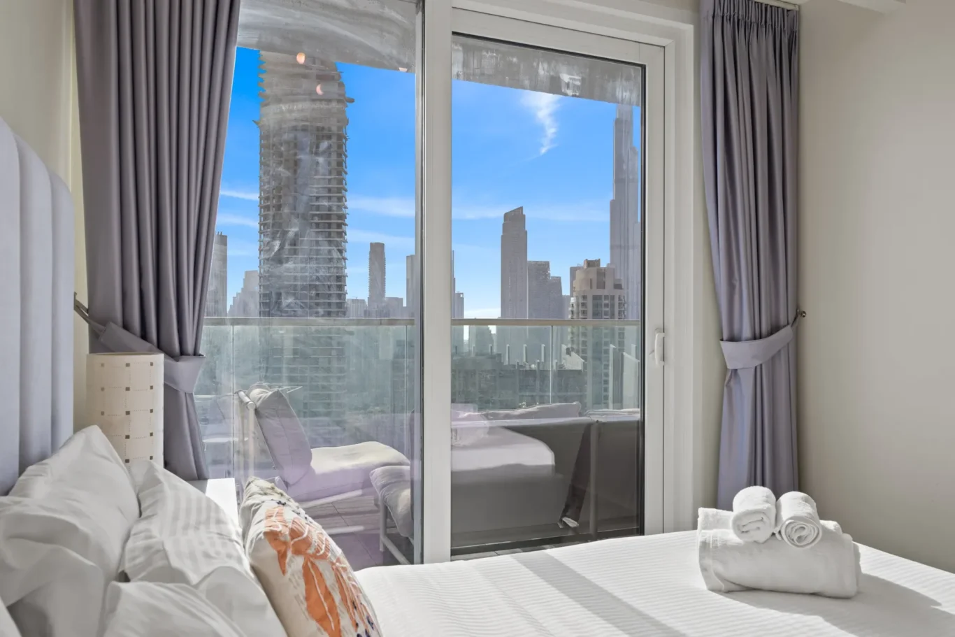 Bedroom with white linens and decorative pillows, glass doors to a balcony overlooking a city skyline with blue sky,; rolled towels on a dresser in the foreground.