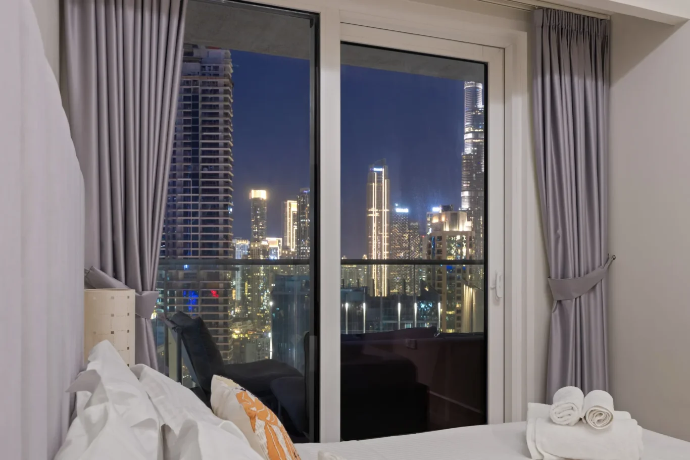 Hotel room with a nighttime city skyline visible through large glass doors; gray curtains frame the view and white bedding is in the foreground.