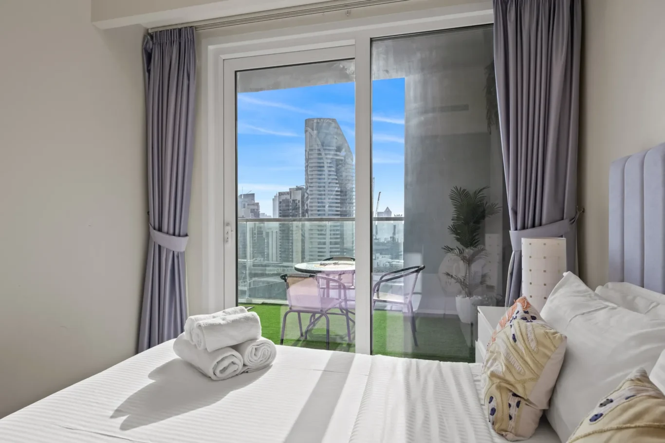 Hotel bedroom with white bed, rolled towels, and a sliding glass door to a balcony; city buildings visible outside against a blue sky.