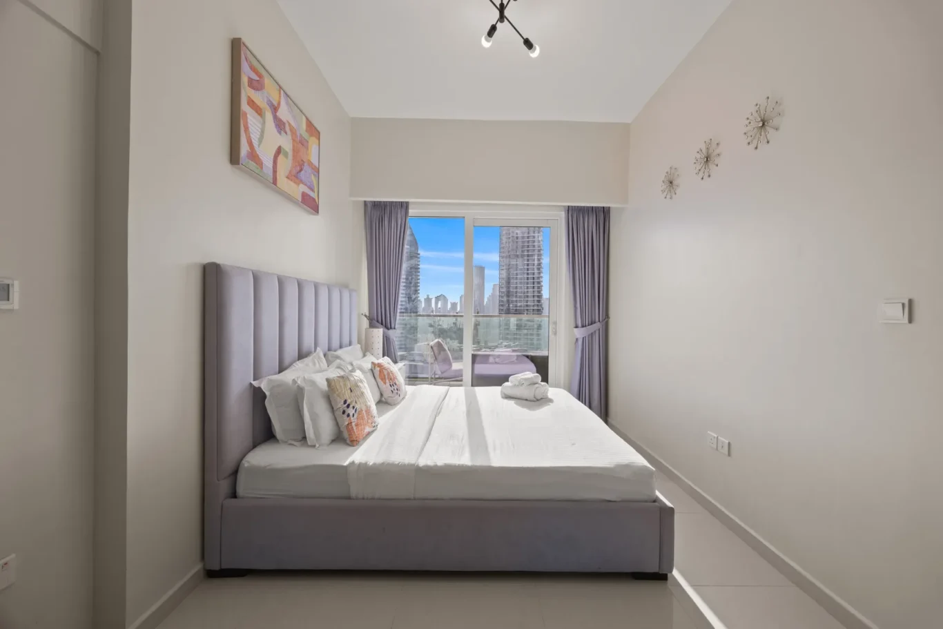 Modern hotel bedroom with a gray upholstered bed, white sheets, and folded towels on the bed; balcony window shows city skyline outside.