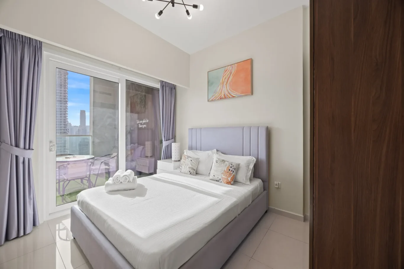 Hotel-style bedroom with gray upholstered bed, white bedding, and decorative pillows; sliding glass doors to a balcony with purple curtains and city view.