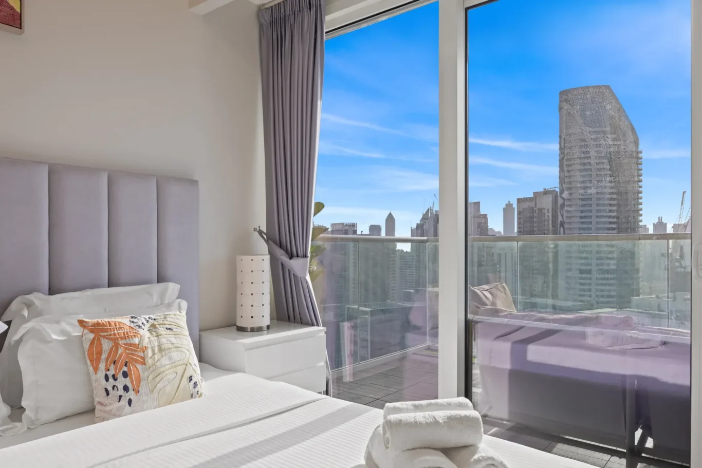 Modern hotel bedroom with a plush gray headboard, white bedding, and a nightstand featuring a cylindrical lamp, overlooking a city skyline through floor-to-ceiling glass doors and balcony railing.