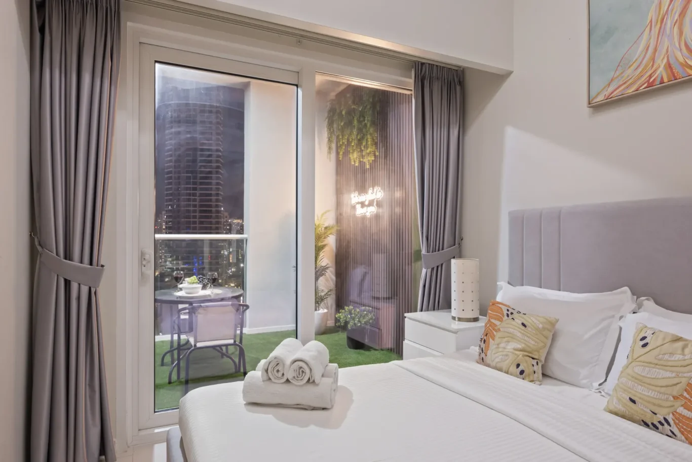 Bedroom with white bedding and decorative pillows, gray headboard, and a balcony door opening to a city view.