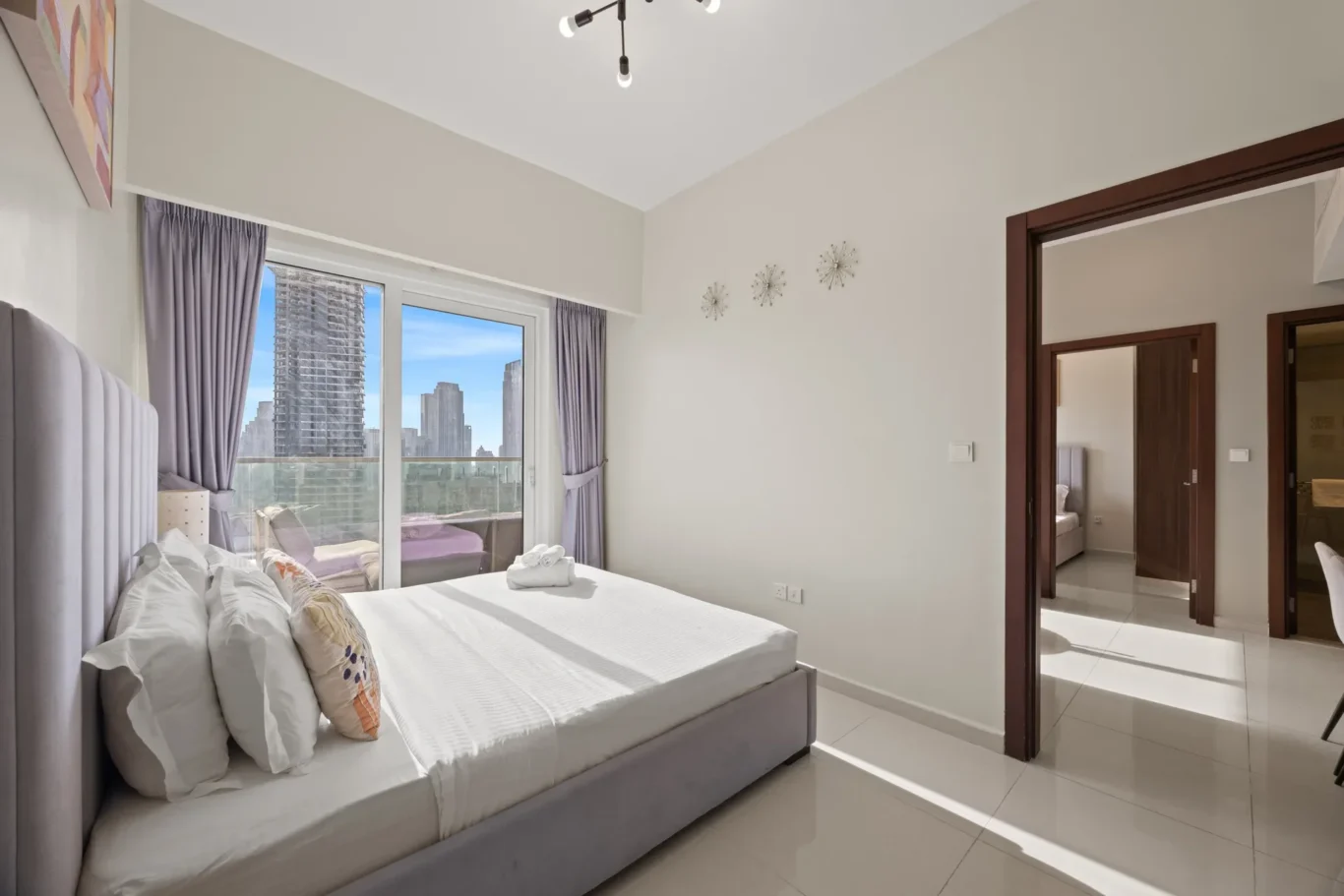 Hotel bedroom with a large white bed and gray headboard, purple curtains, and a balcony overlook of city skyscrapers.
