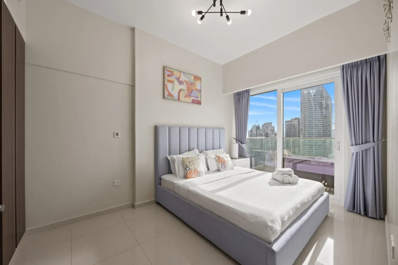 Hotel bedroom with a gray upholstered bed, white bedding, and decorative pillows, facing a large window with a city skyline view.