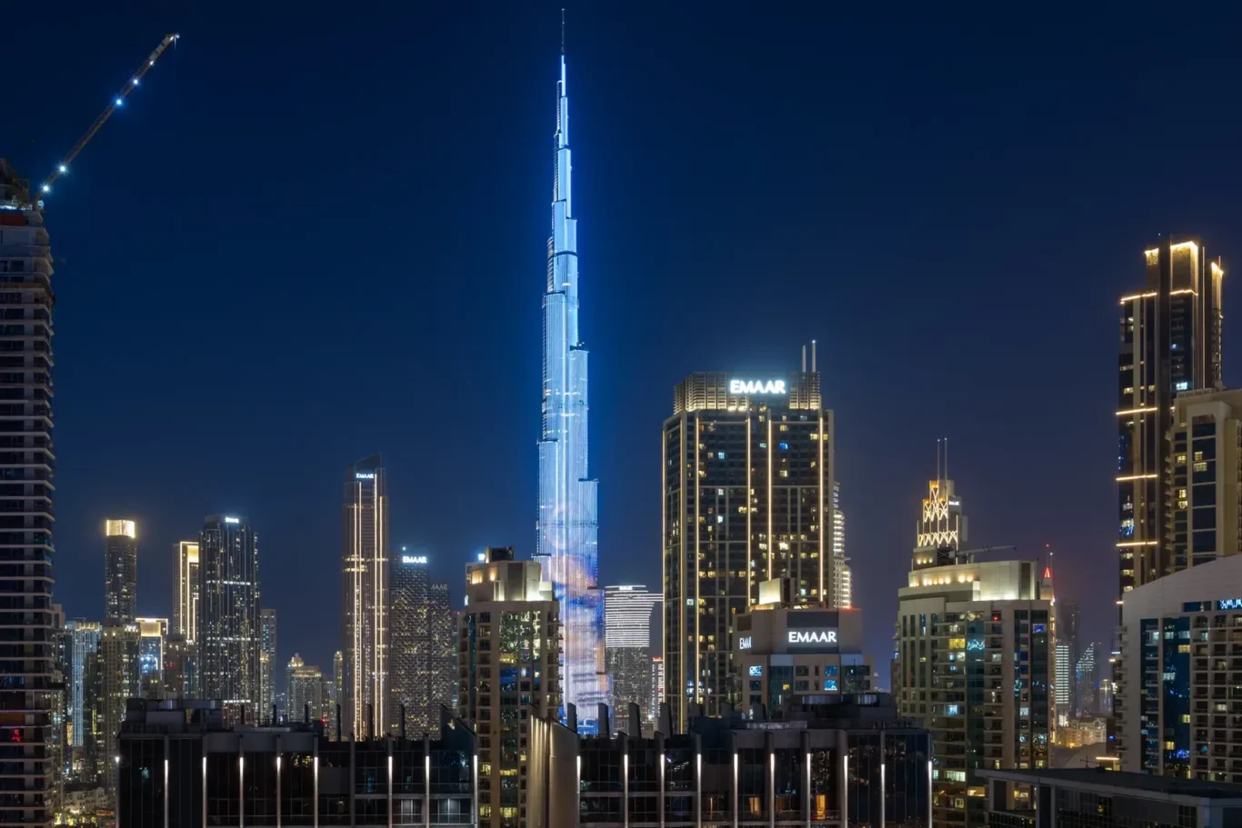 Night skyline of Dubai with the blue-lit Burj Khalifa towering above surrounding skyscrapers.