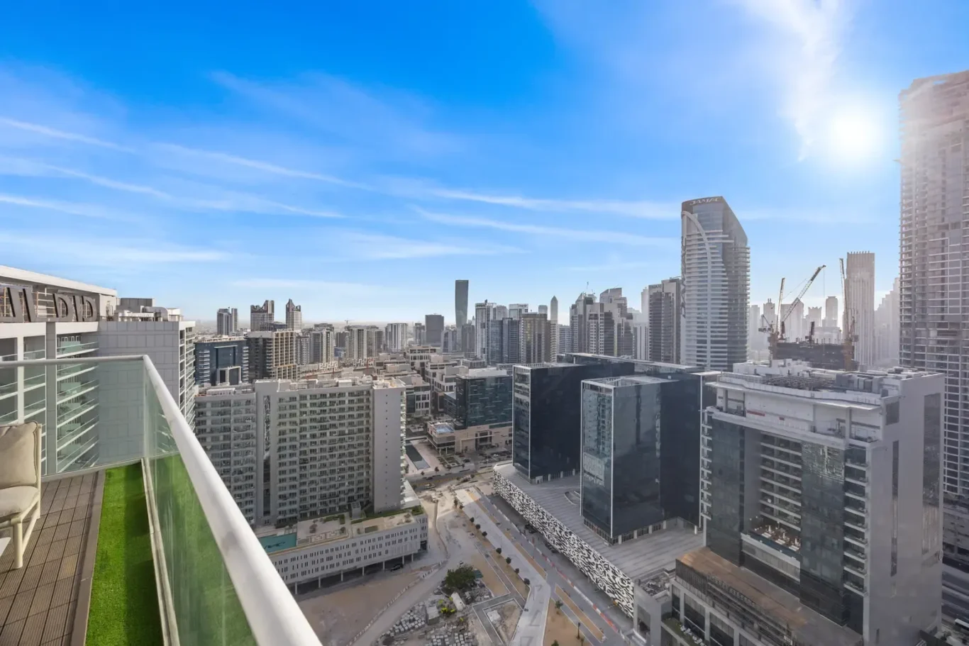 Balcony railing and chair overlook a dense city skyline of modern high-rise buildings under a bright blue sky with the sun to the right.