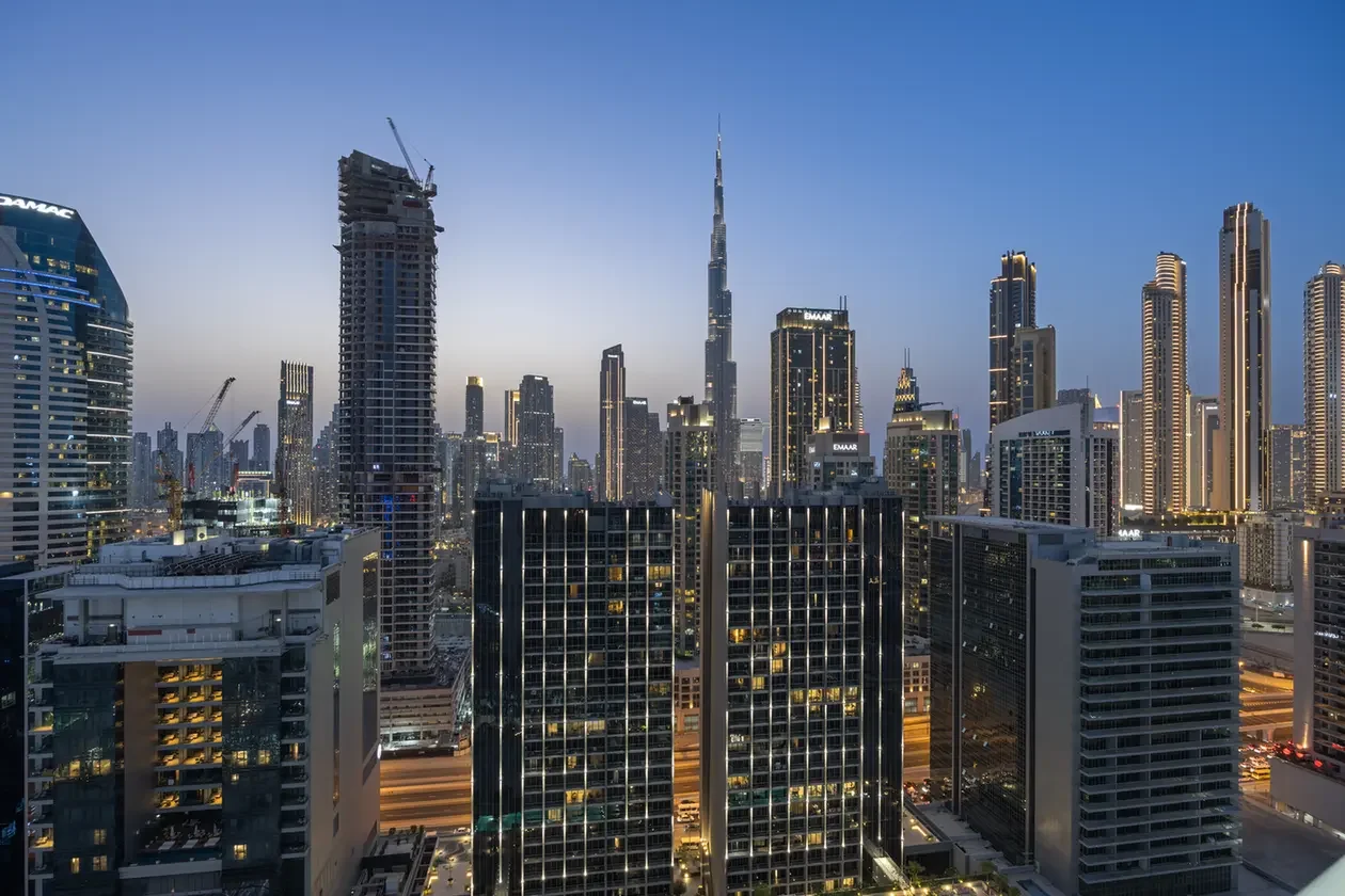 Dubai skyline at dusk with a cluster of illuminated skyscrapers and a tall central tower against a clear blue sky.