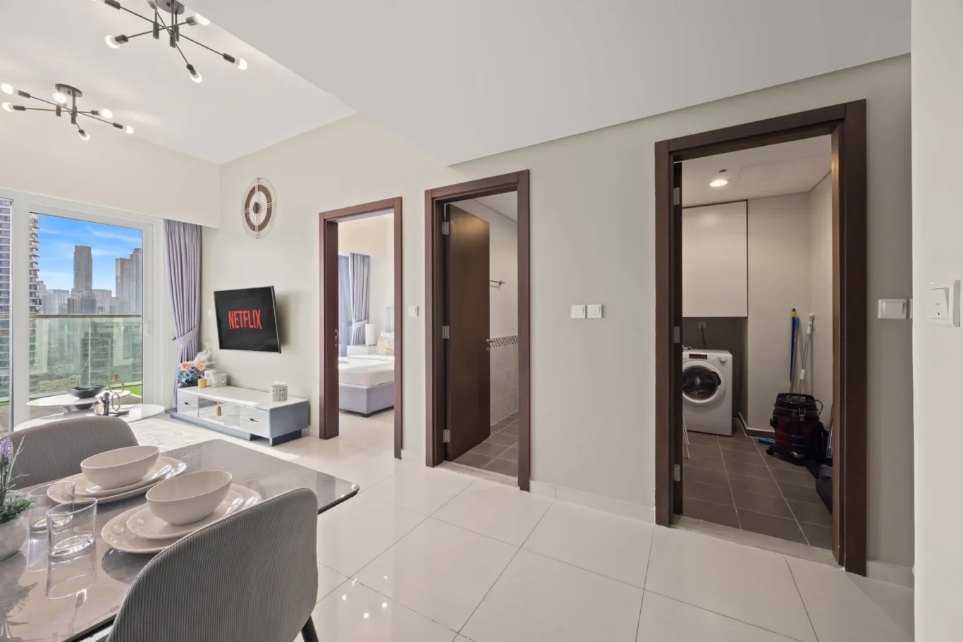 Open-plan living area with a dining table, wall-mounted Netflix TV, and two doorways to bedrooms; balcony with city skyline on the left.