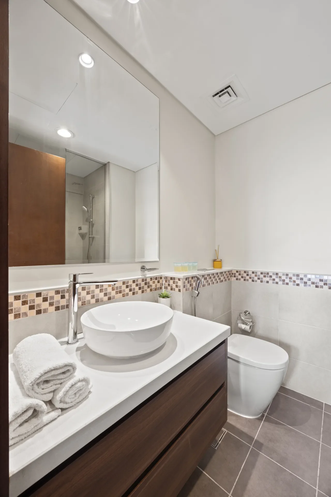 Modern bathroom featuring a white vessel sink on a white countertop, a dark wood vanity, a large mirror, and a tiled floor; toilet in the background with a shower enclosure visible beyond.