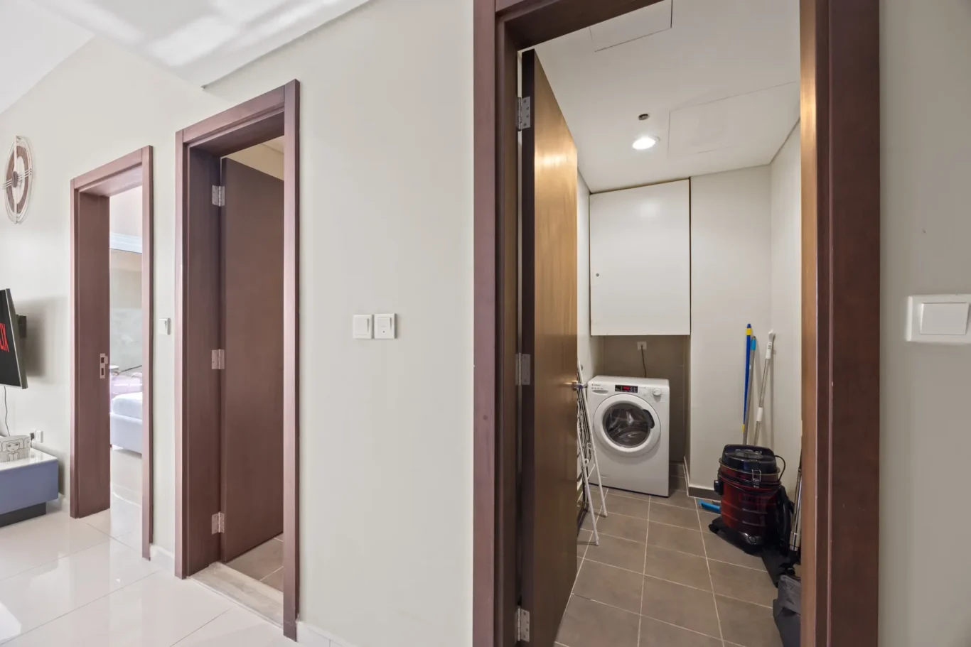 Narrow hallway with open doors to a laundry room containing a front-loading washing machine and cleaning supplies.