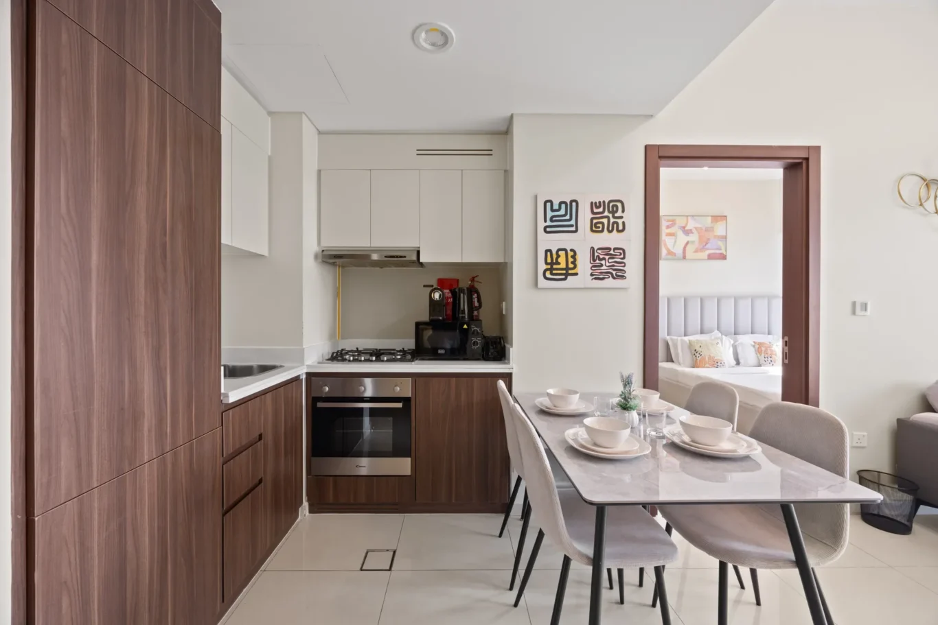 Modern kitchen with dark wood cabinets, white countertops, stainless oven and microwave, and a marble dining table set for six; doorway to a bedroom visible nearby.