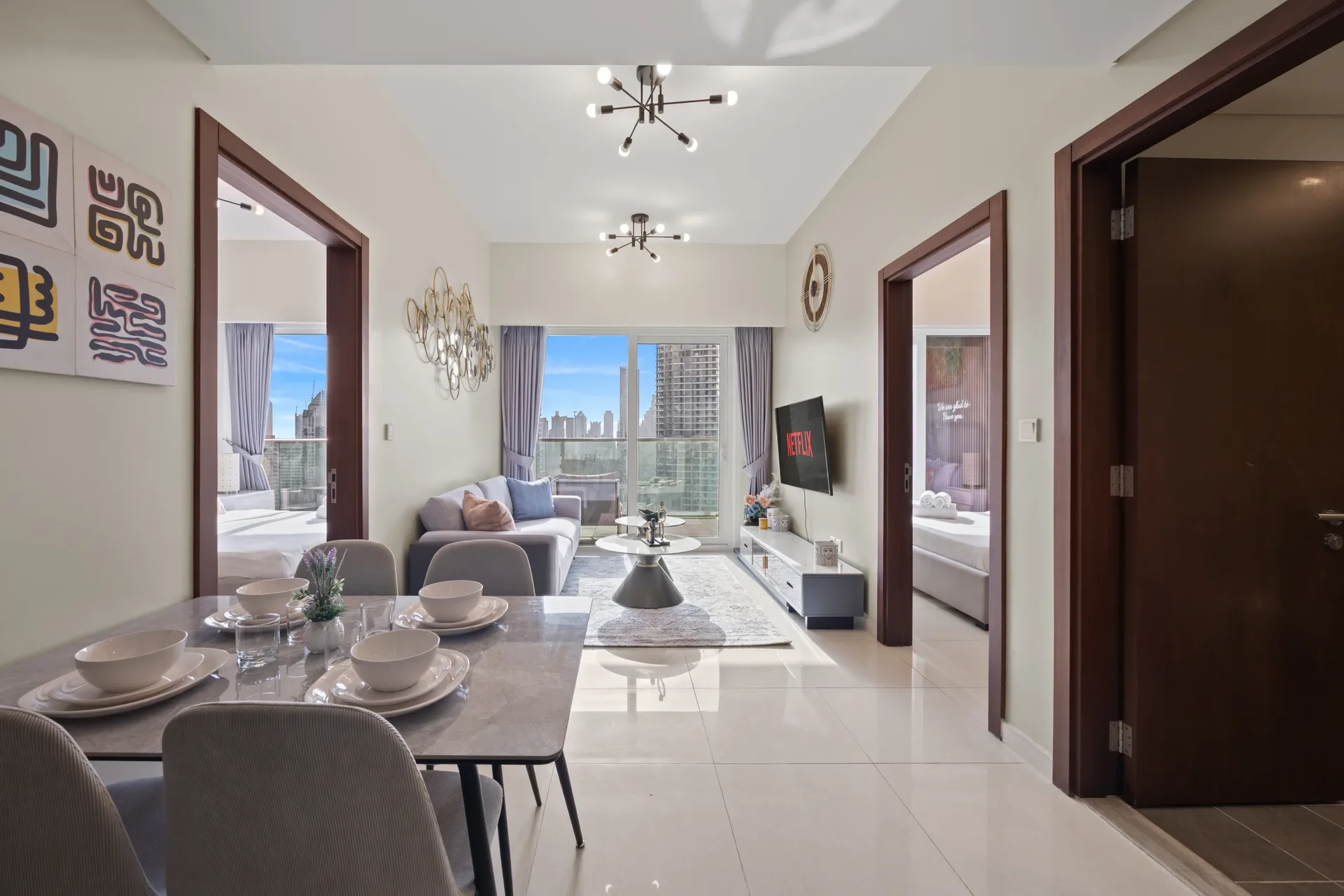 Dining area with a marble table set for six, gray chairs, and a small plant centerpiece; open living room beyond with a sofa and coffee table, city skyline visible through large windows.