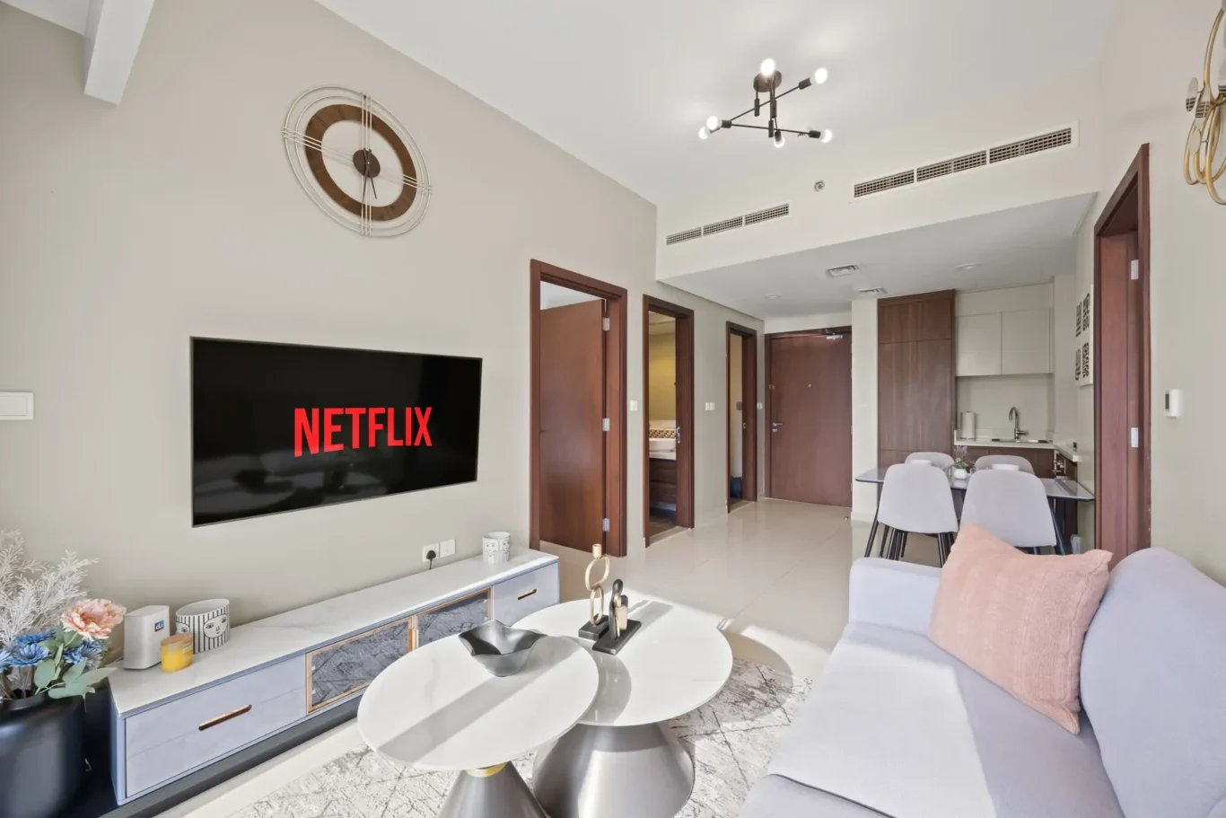 Living room with a wall-mounted flat-screen Netflix TV, a light-grey sofa with a pale pink cushion, and round white coffee tables.