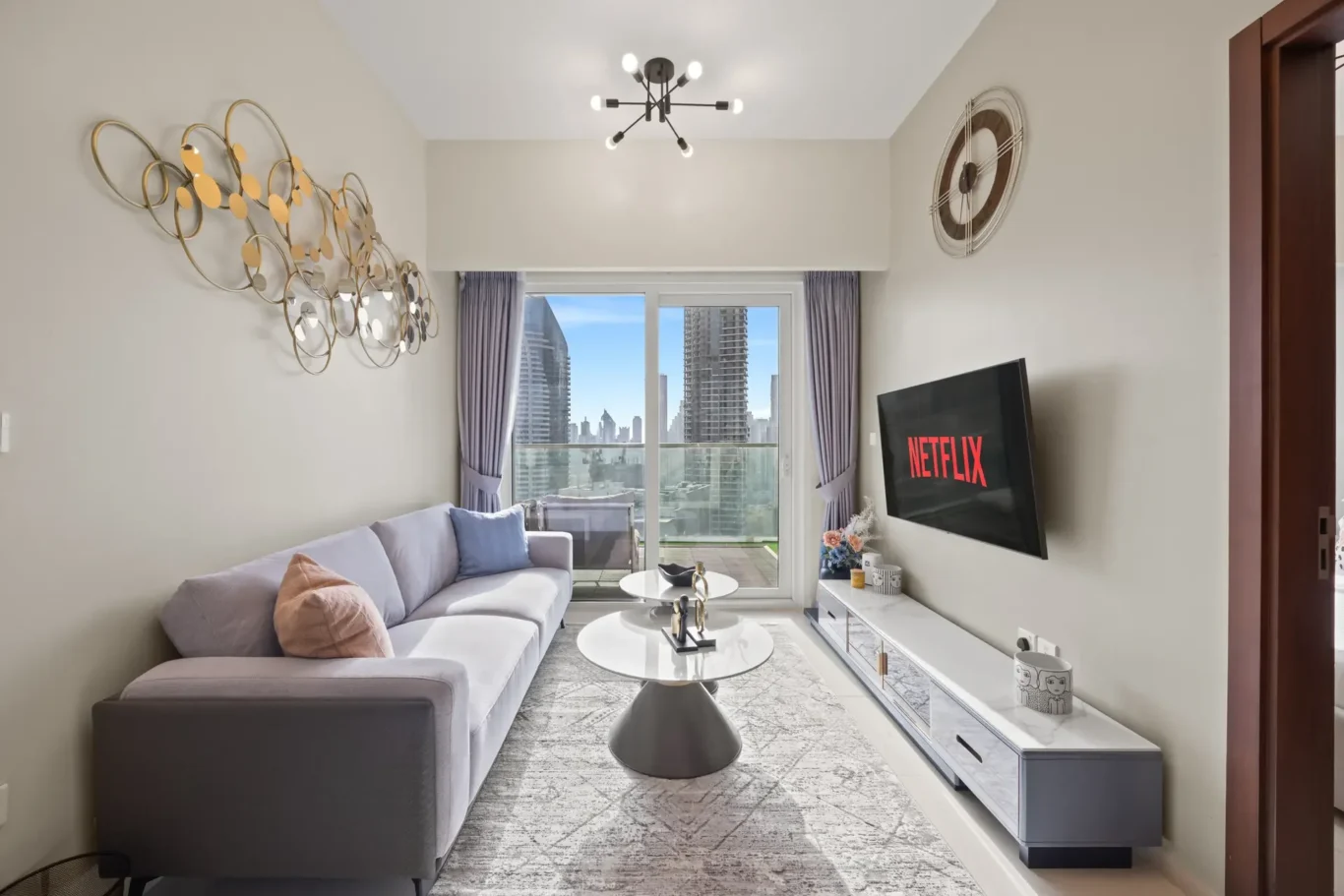 Modern living room with gray sectional, gold circular wall sculpture, and a wall-mounted Netflix TV; city skyline visible through balcony doors.