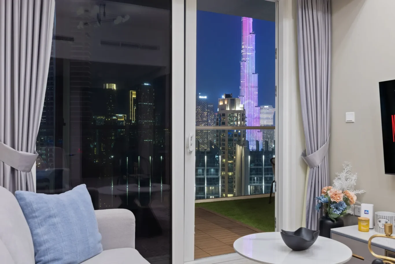 Living room at night with glass doors opening to a balcony; city skyline visible, including a tall purple-lit skyscraper.