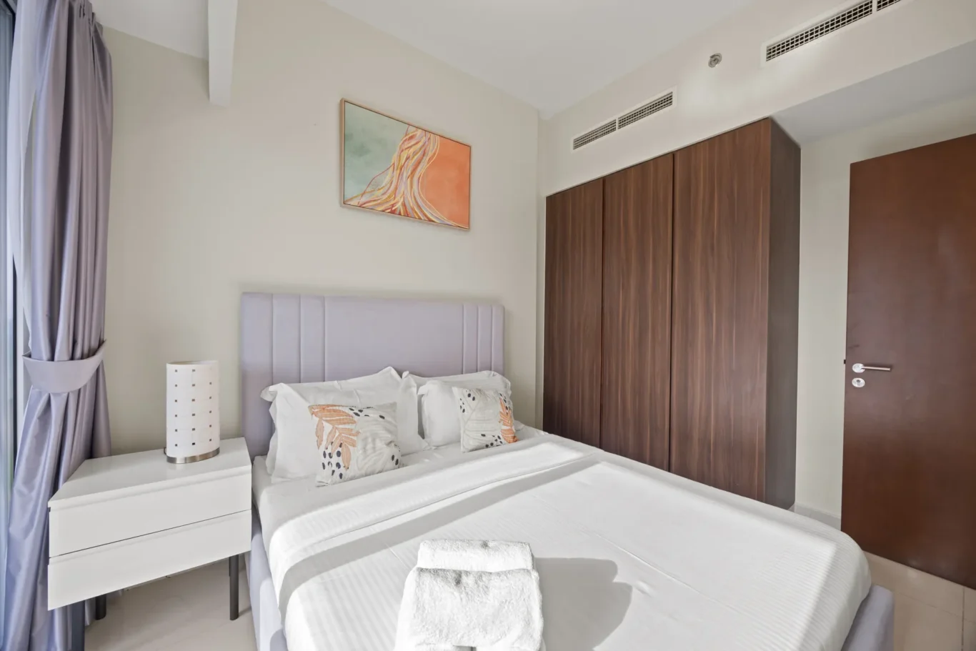 Hotel bedroom with two white-pillowed beds, gray headboard, and a tall white nightstand beside a lavender-curtained window.