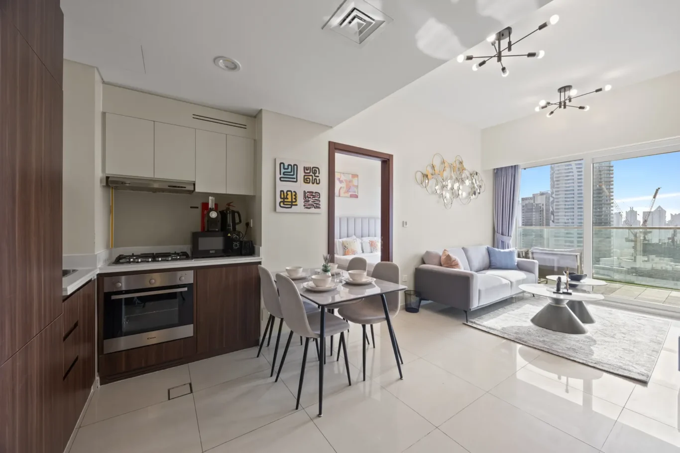 Open-plan living area with a wood-accent kitchen on the left, a glass dining table, gray chairs, and a sofa by large balcony windows overlooking the cityscape.