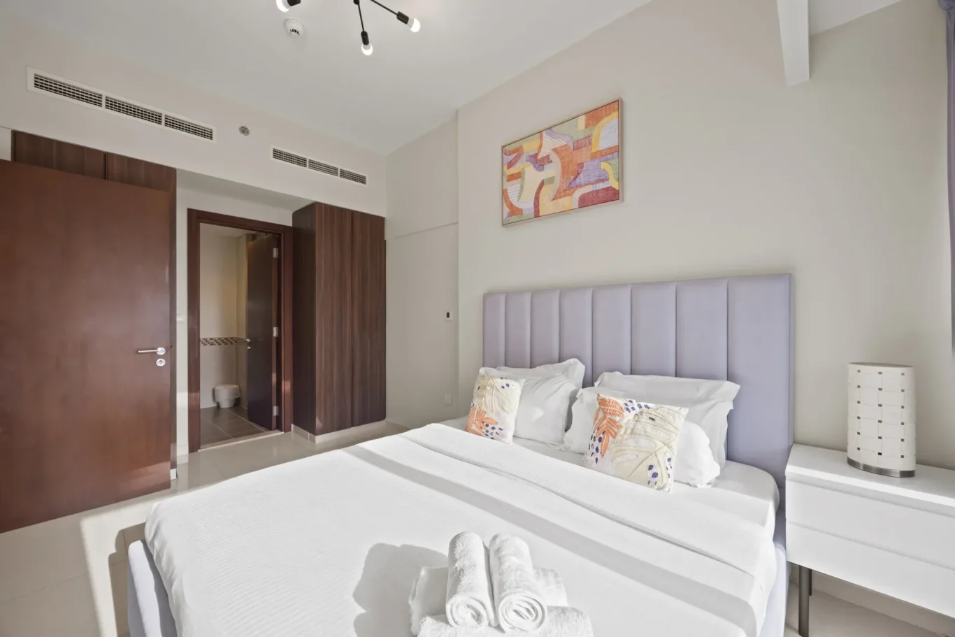 Double bed with white linens and lavender headboard in a bright hotel room, decorative pillows on the bed, and a nightstand with a lamp nearby.