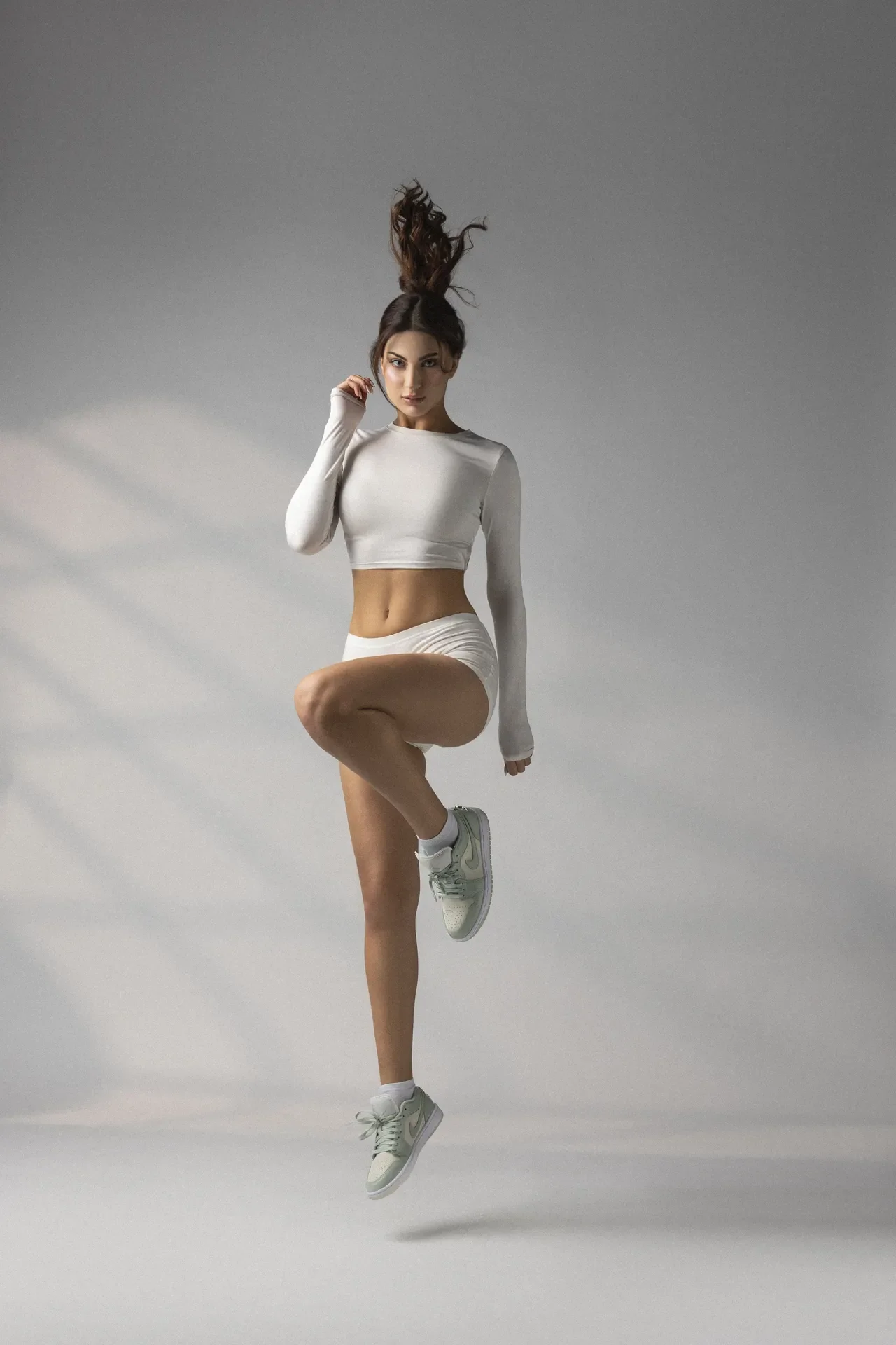 Athlete in a white crop top and shorts jumping mid-air against a gray studio backdrop with flowing hair.