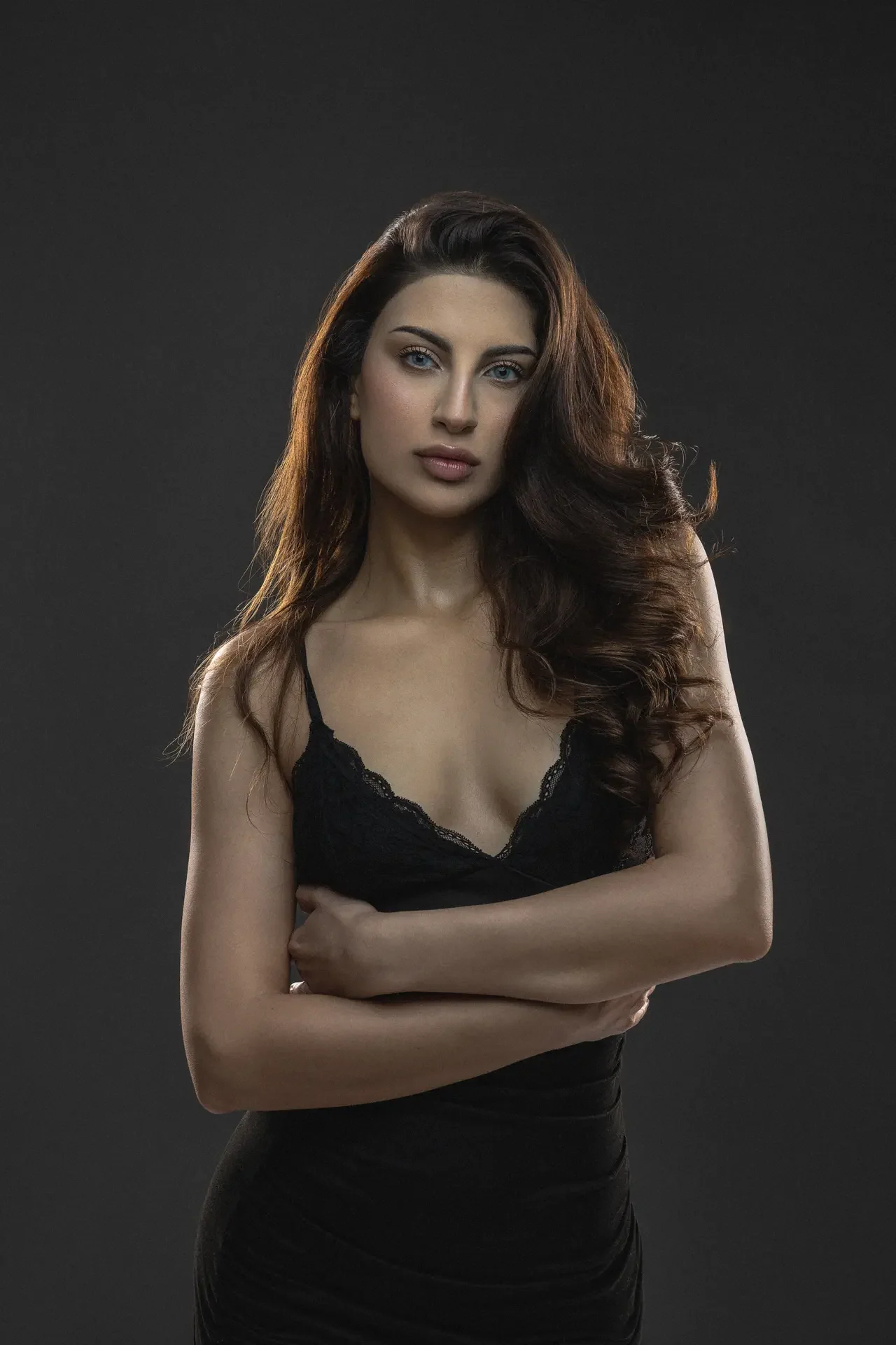 Portrait of a woman with long wavy brown hair wearing a black lace top, arms crossed, against a dark background.