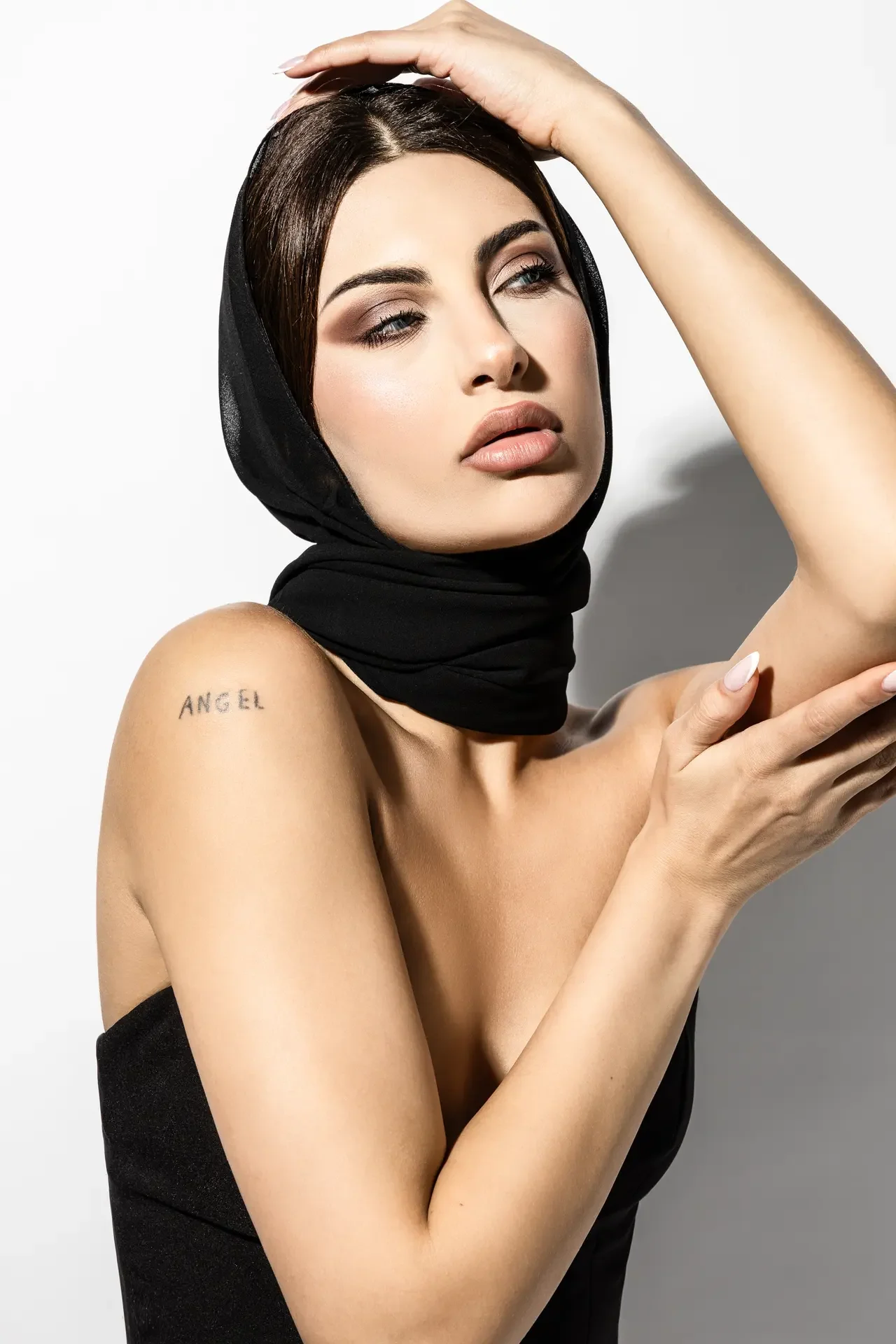 Woman wearing a black headscarf, neutral makeup, and a visible tattoo reading ANGEL on her upper arm, resting her hand on her head.