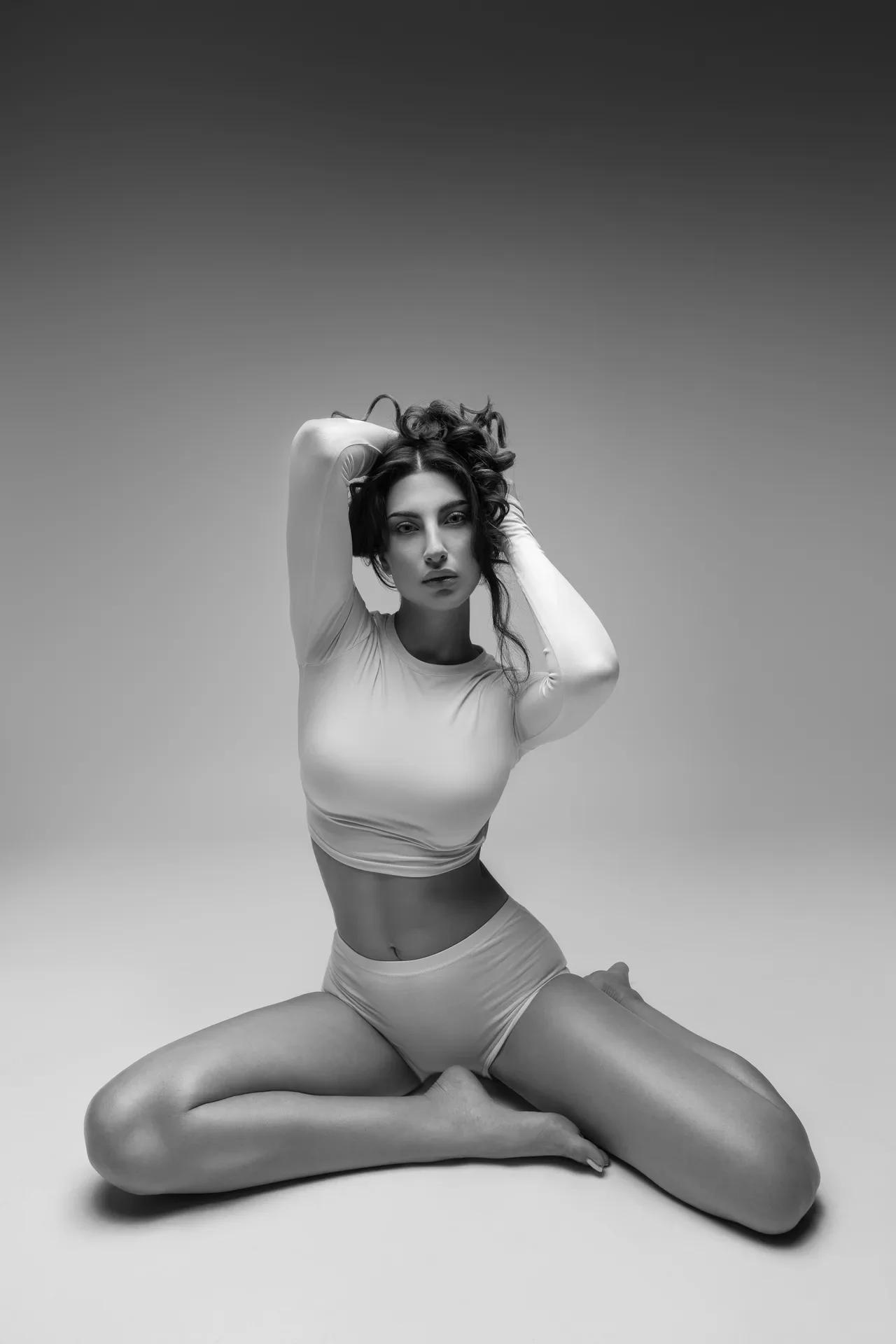 Black-and-white photo of a woman in athletic wear seated cross-legged, arms raised behind her head, looking at the camera in a studio.