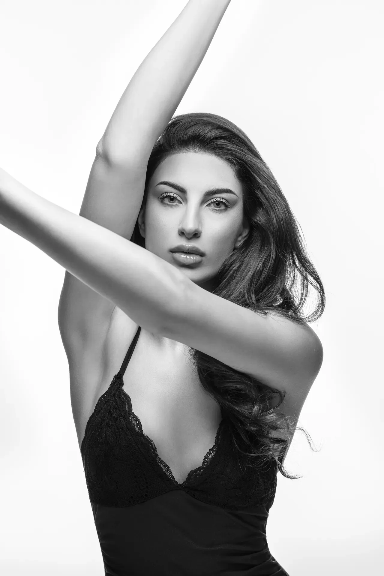 Black-and-white portrait of a woman with long hair in a lace black top, arms raised above her head, looking at the camera.