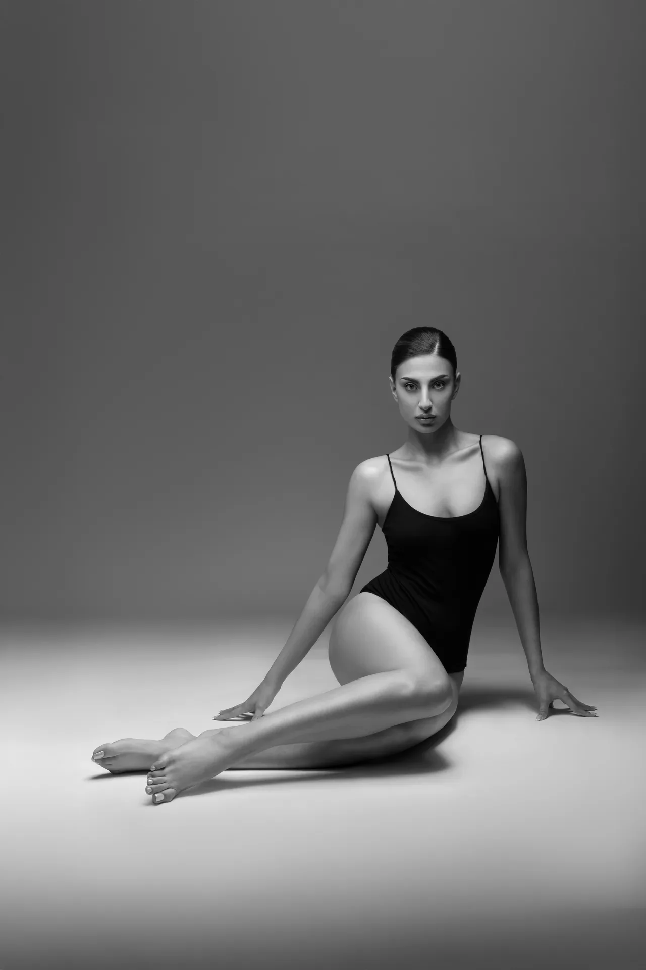 Black-and-white photo of a woman in a black one-piece swimsuit seated on the floor with legs extended, looking at the camera.