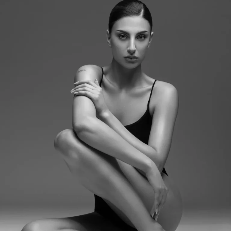 Black-and-white portrait of a woman in a black leotard seated with legs crossed and arms wrapped around her knee, looking at the camera (tattoo on her left arm).