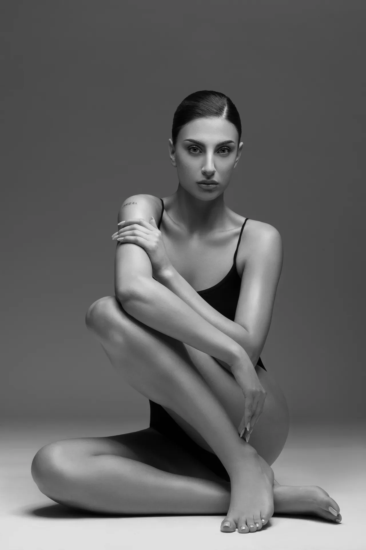 Black-and-white portrait of a woman in a black leotard seated with legs crossed and arms wrapped around her knee, looking at the camera (tattoo on her left arm).