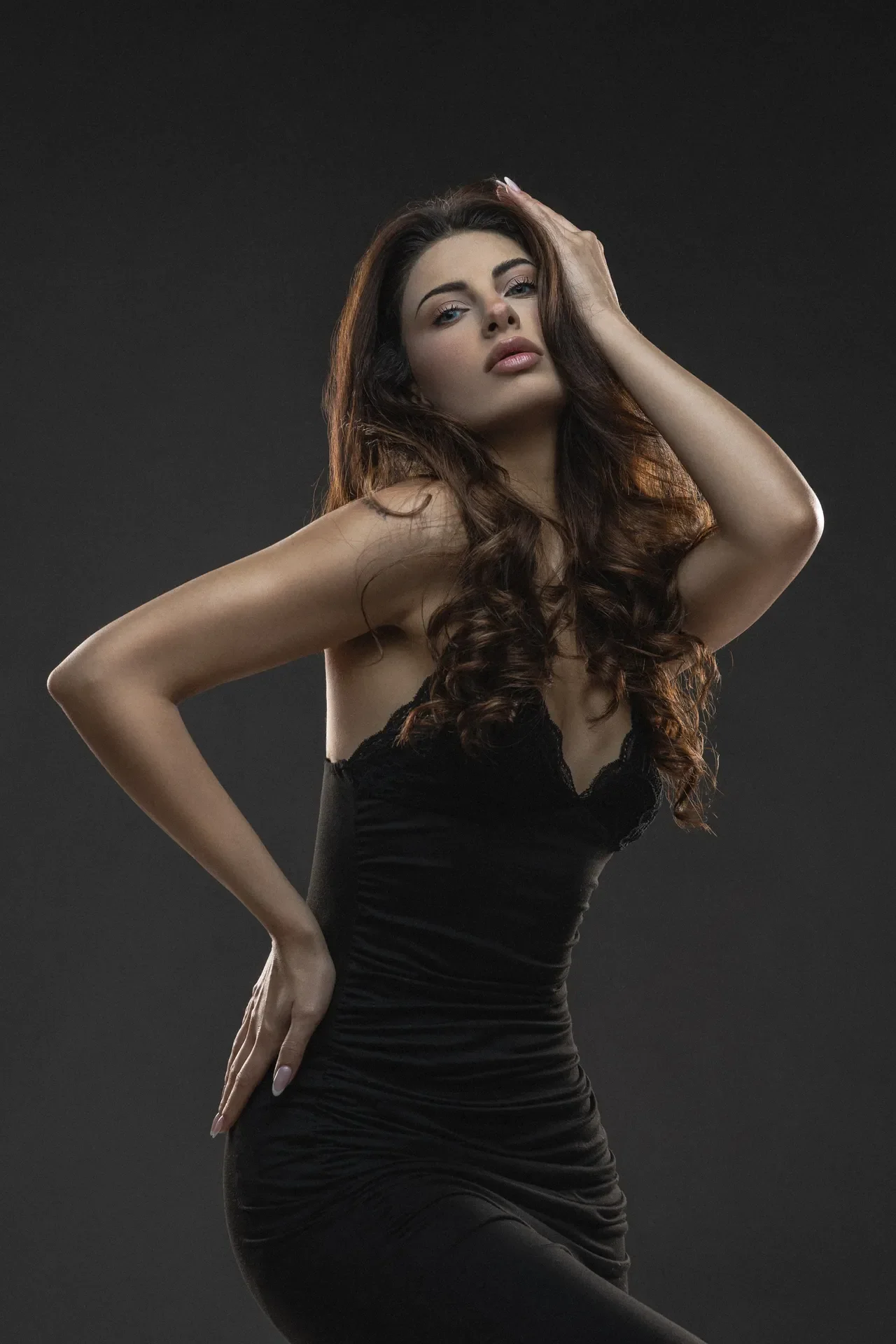 Woman posing in a black dress with one hand on her head against a dark backdrop in a studio setting.