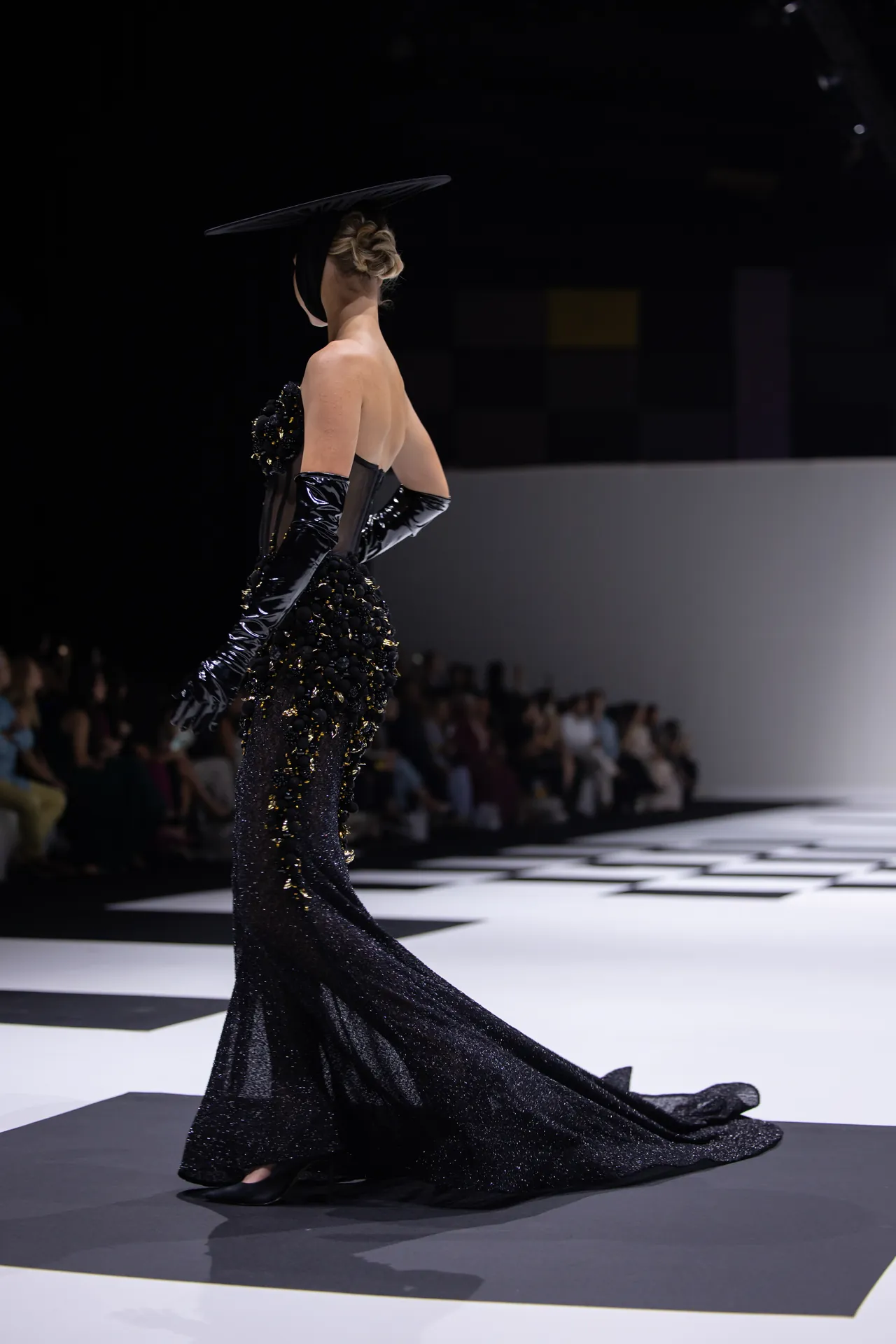 Model on a runway wearing a floor-length black sequined gown with long gloves and a dramatic headpiece, spotlight on her as audience watches.