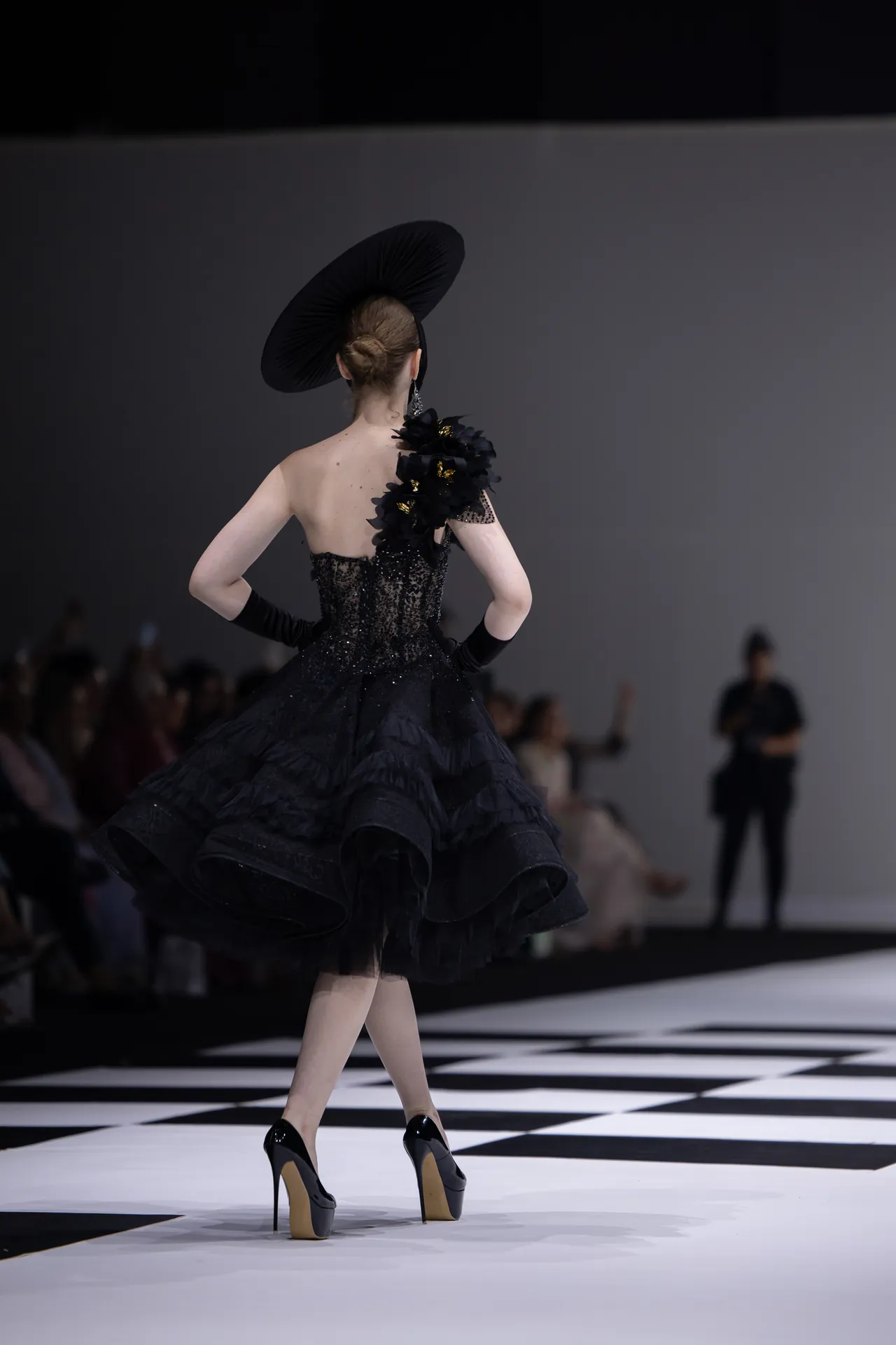 Back view of a model on a black-and-white checkered runway wearing a voluminous black gown and a large circular hat, audience blurred in the background.