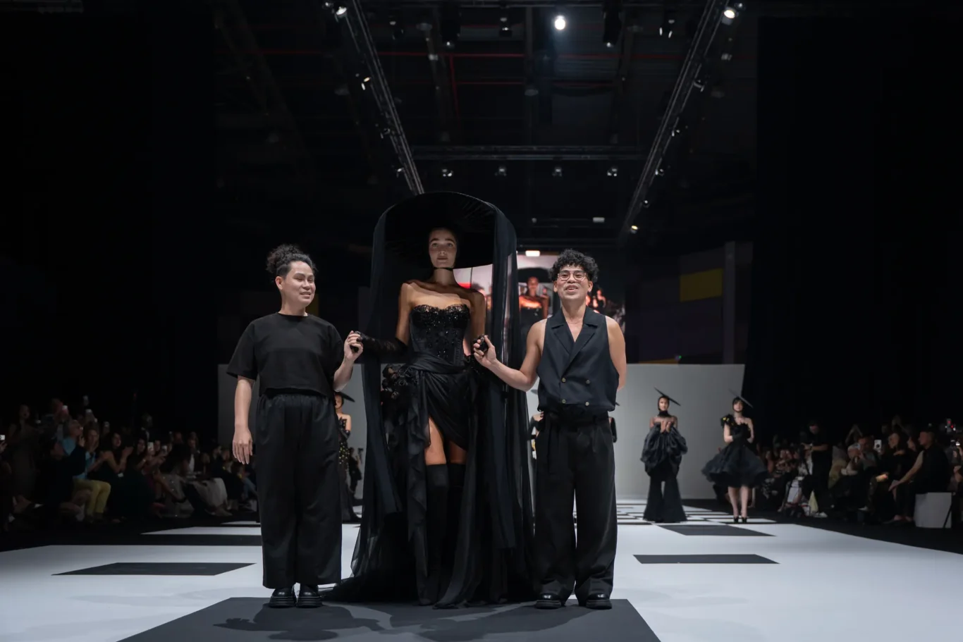 Models on a fashion runway; central figure wears a black corseted gown with a cape, flanked by two assistants.