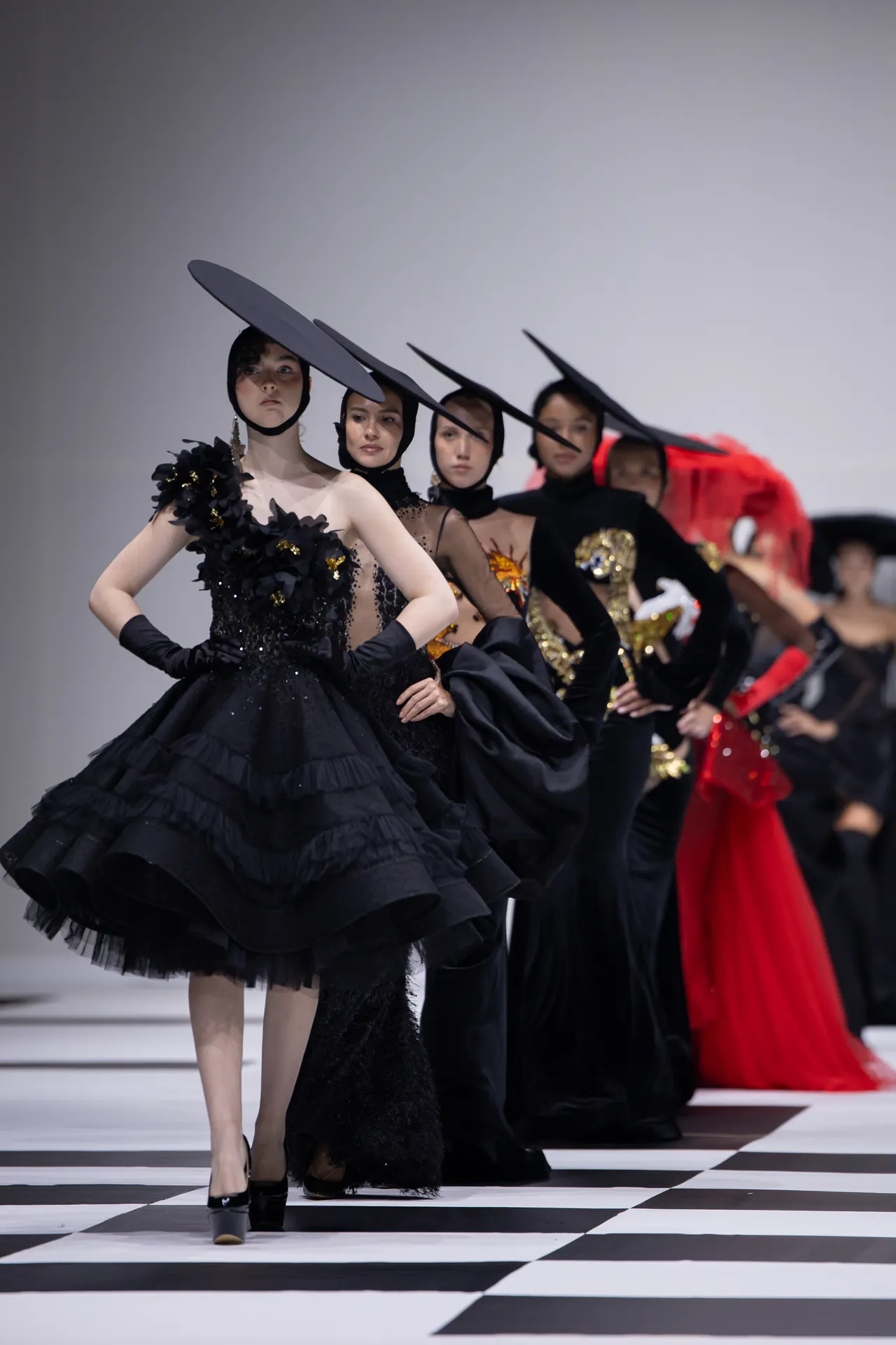 Models in black gowns with oversized hats pose in a runway line on a black-and-white checkered floor, at a fashion show.
