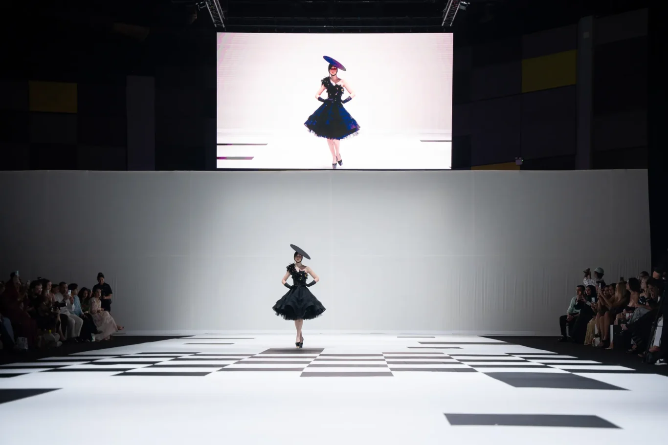 Model in a black, ruffled cocktail dress and wide hat walks a black-and-white checkerboard runway, with spectators seated along both sides and a large screen backdrop behind.
