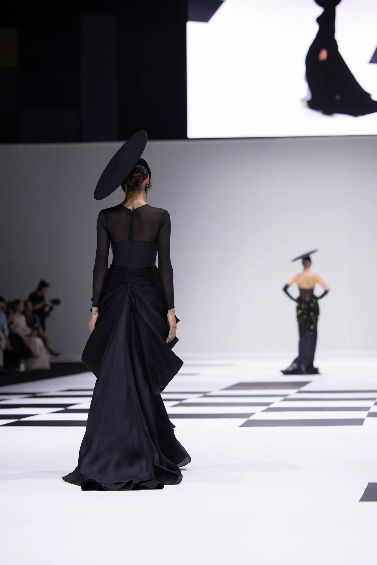 Back view of a model walking a runway in a long black gown with a large circular black hat; checkerboard floor visible in foreground.