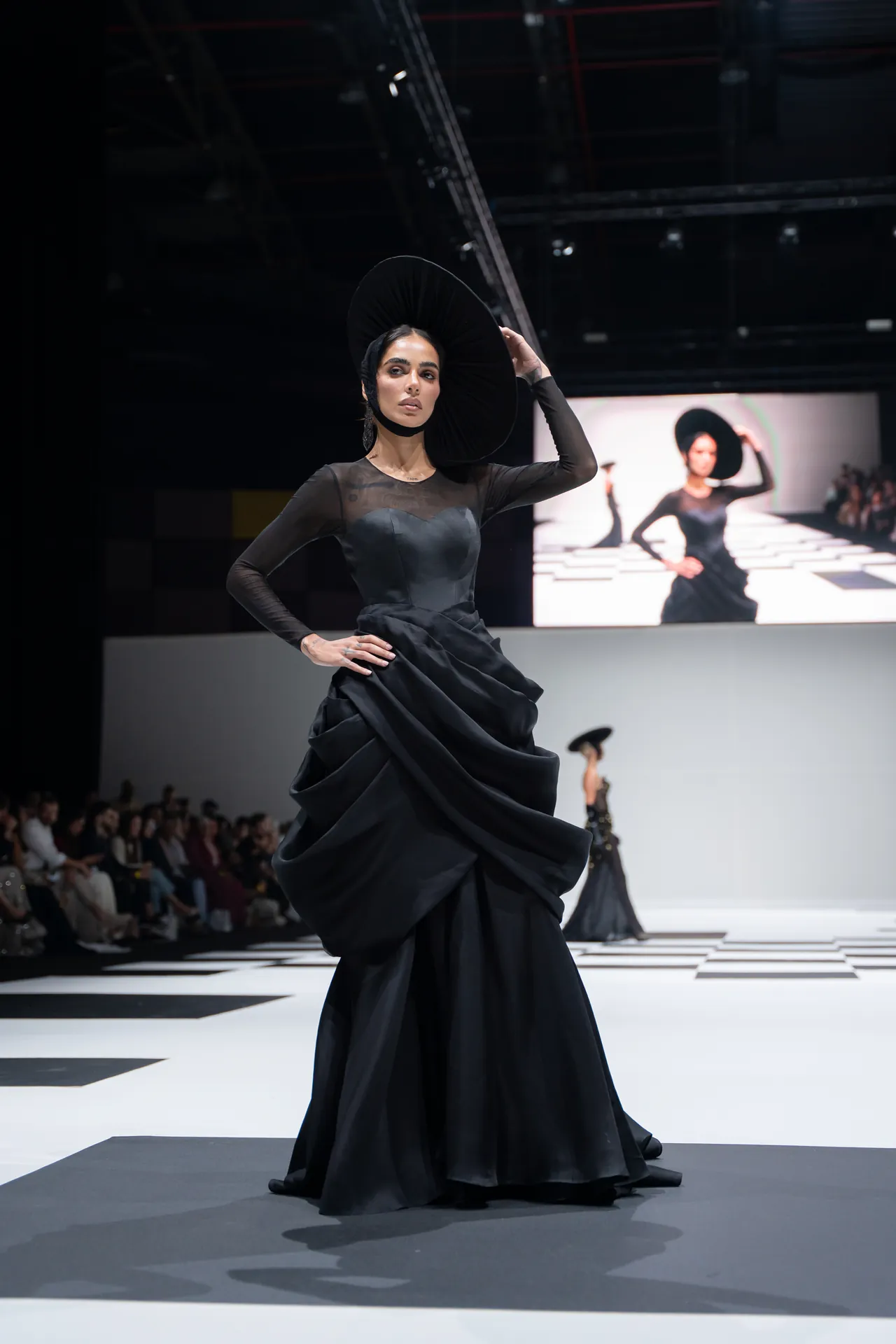 Model on a runway wearing a black, voluminous gown and a large circular hat, striking a pose under bright lights.