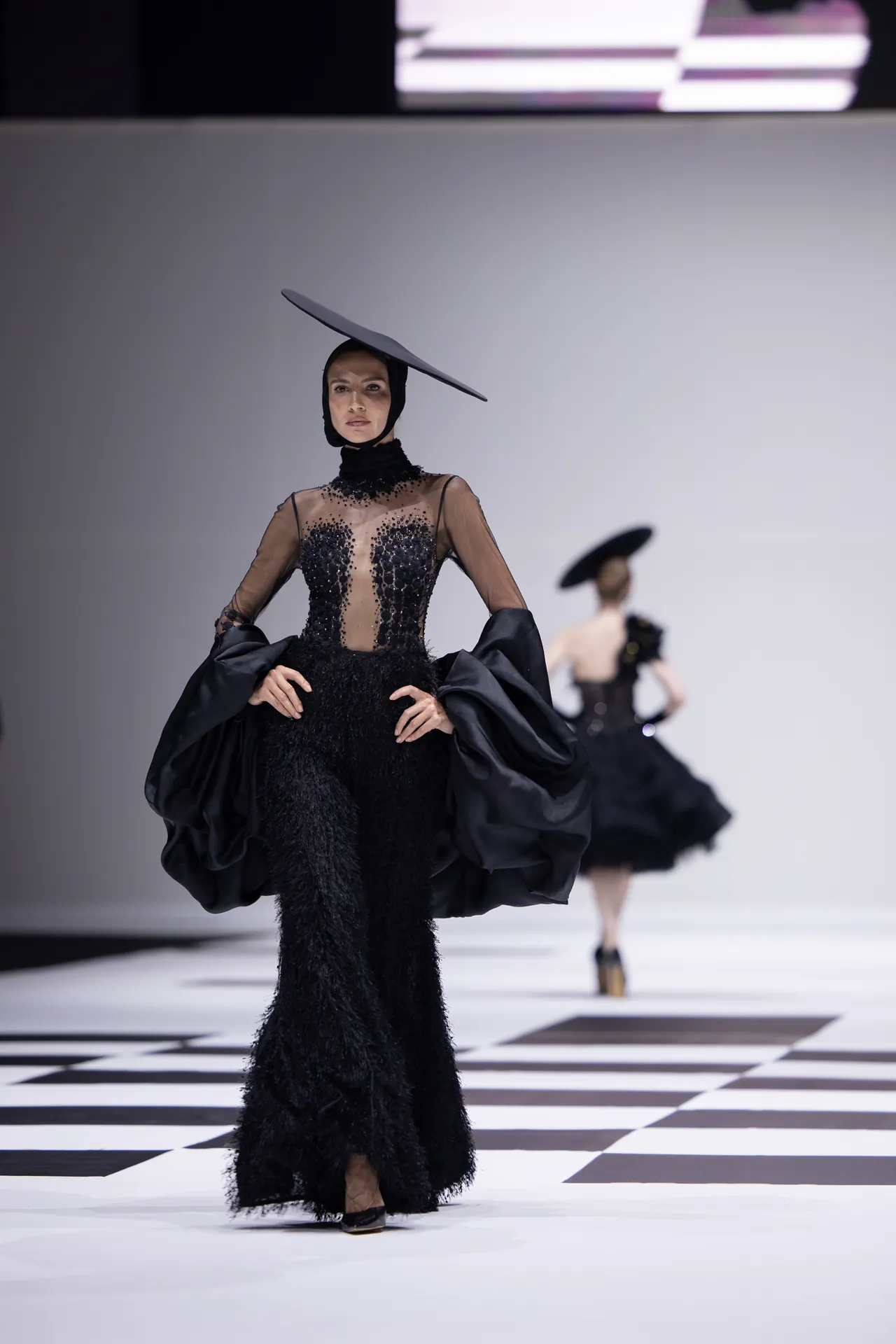 Model on a fashion runway wearing a black sheer-beaded gown with oversized cape sleeves and a tilted hat.