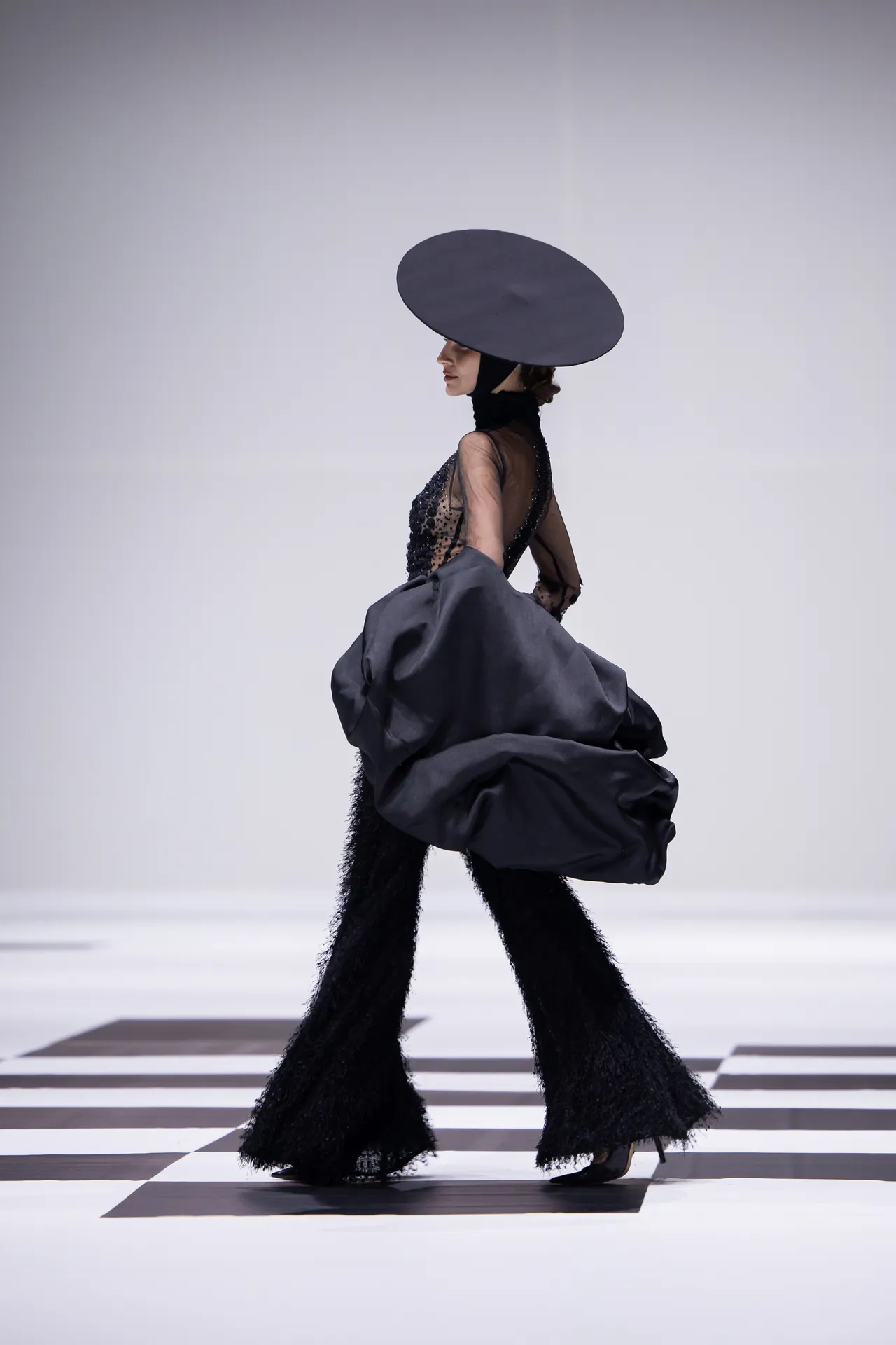 Model walking on a runway in a dramatic black ensemble with a large circular hat and voluminous skirt; geometric checkered floor beneath.