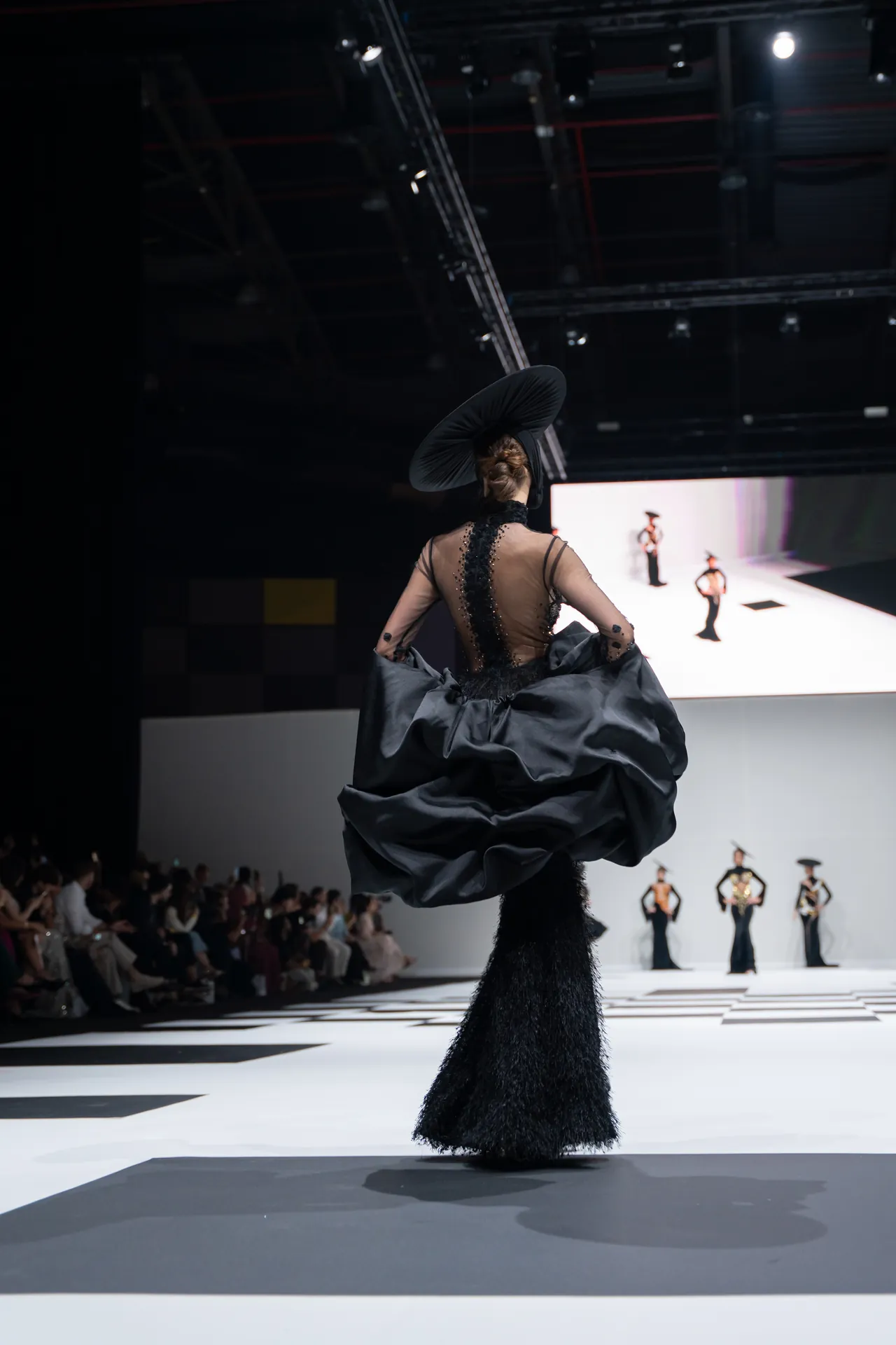Back view of a model on a runway in a dramatic black gown with a voluminous skirt and wide-brimmed hat, audience in the background.